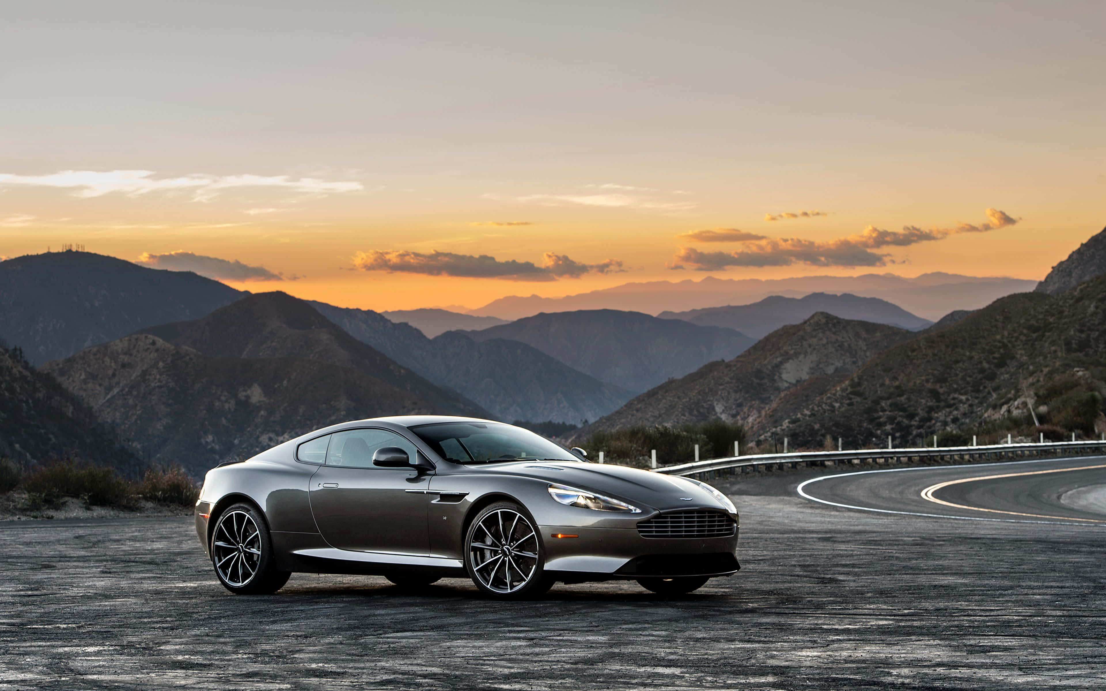 時尚型格嘅Aston Martin DB9 壁紙 — 一架時尚的銀色 Aston Martin 跑車停泊在蜿蜒的山路上，夕陽映照出橙色和紫色的天空。