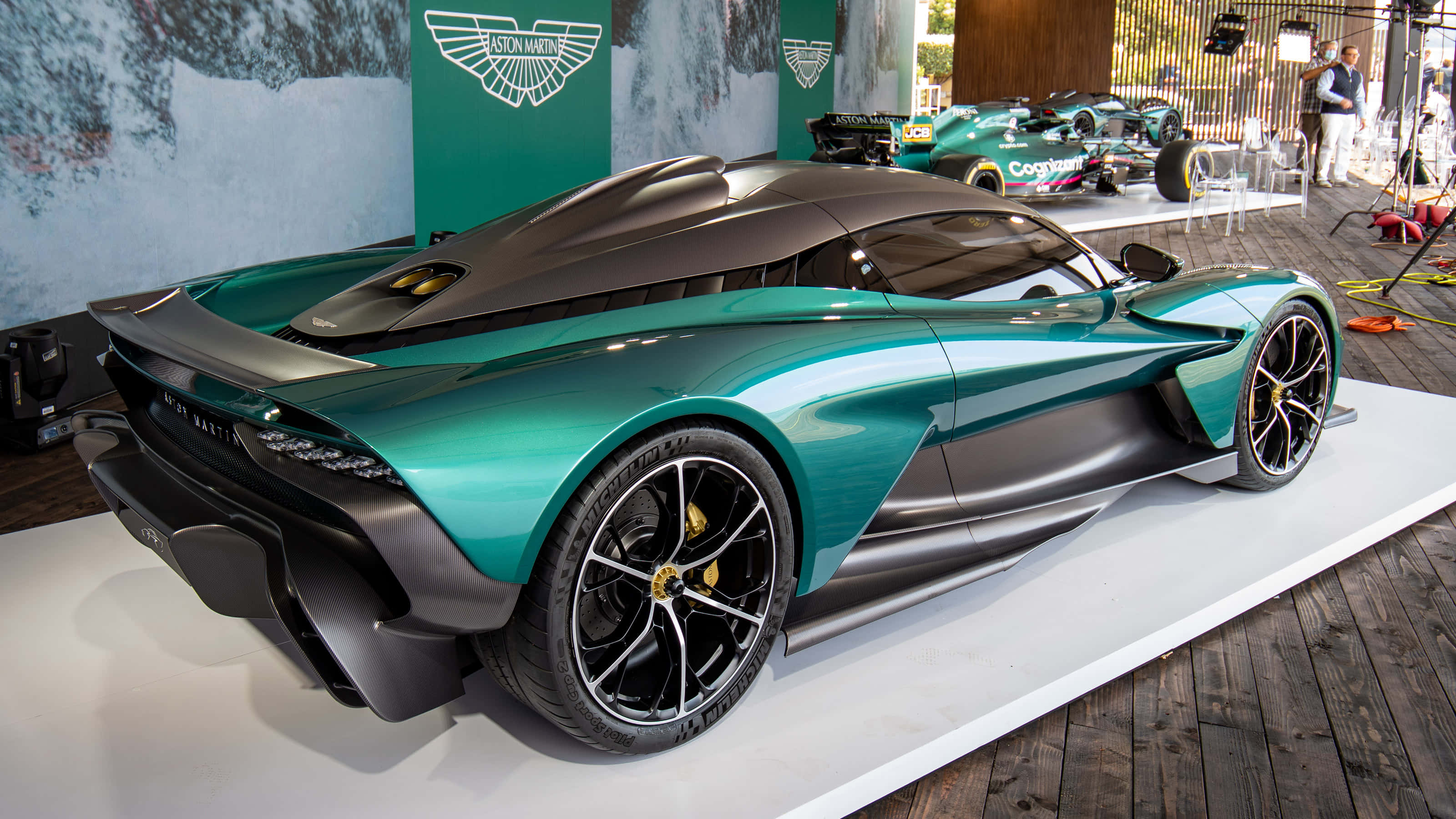 駕駛 Aston Martin，探索新世界 壁紙 — 一輛時尚的藍綠色和黑色Aston Martin hypercar在室內汽車活動中展示在白色平台上。