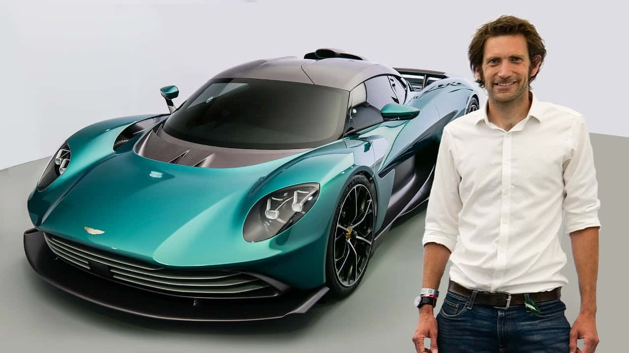 駕馭 Aston Martin，感受駕駛之美 壁紙 — 一名身穿白恤衫的男子站在一輛帶有黑色飾邊的流線型青綠色Aston Martin超跑旁邊，背景為簡約的工作室。