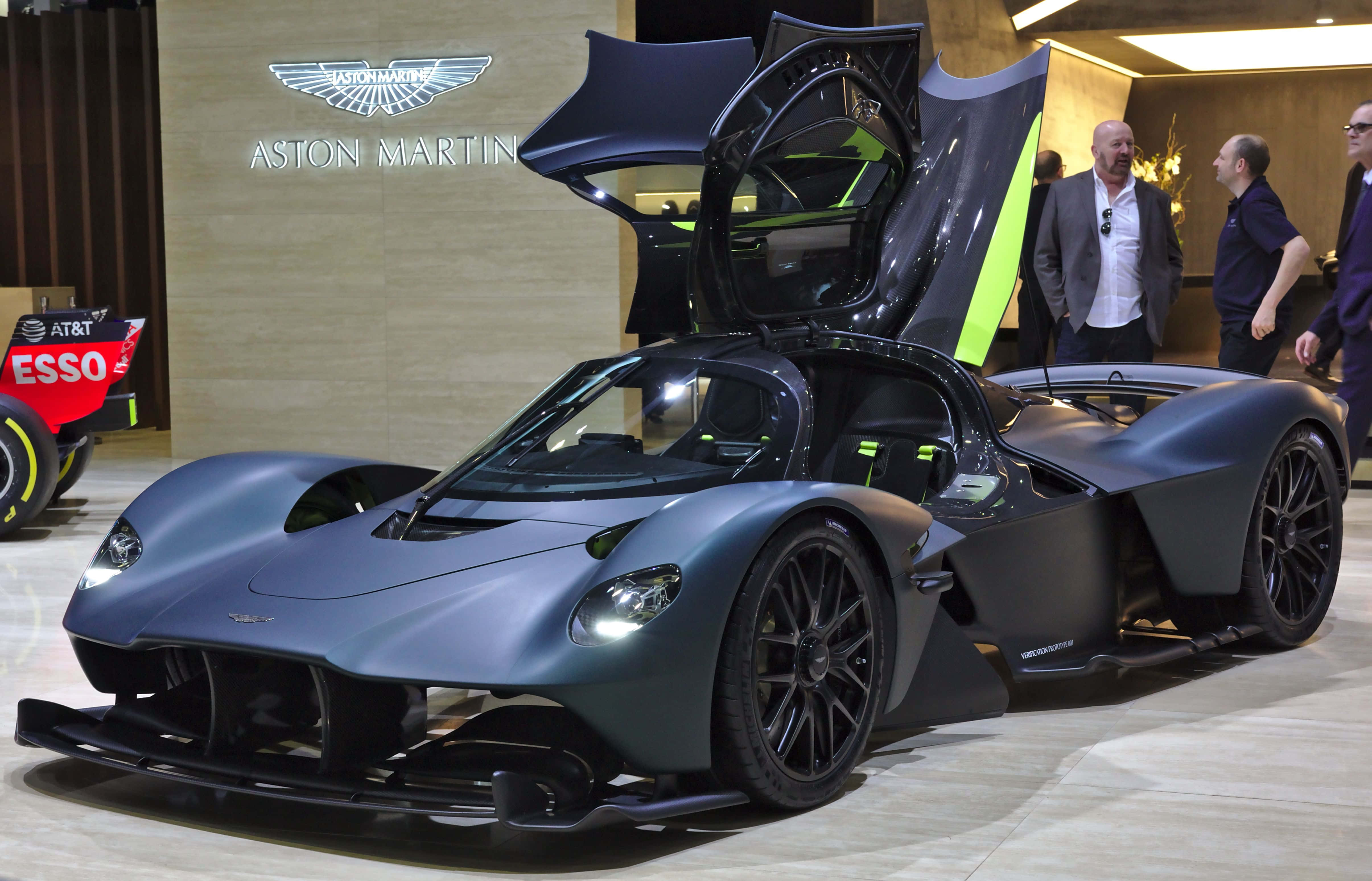 震撼嘅Aston Martin Valkyrie超級跑車全貌 壁紙 — 一輛啞光灰色的Aston Martin超跑，帶有發光的綠色飾邊和打開的鷗翼門，在車展上展出。