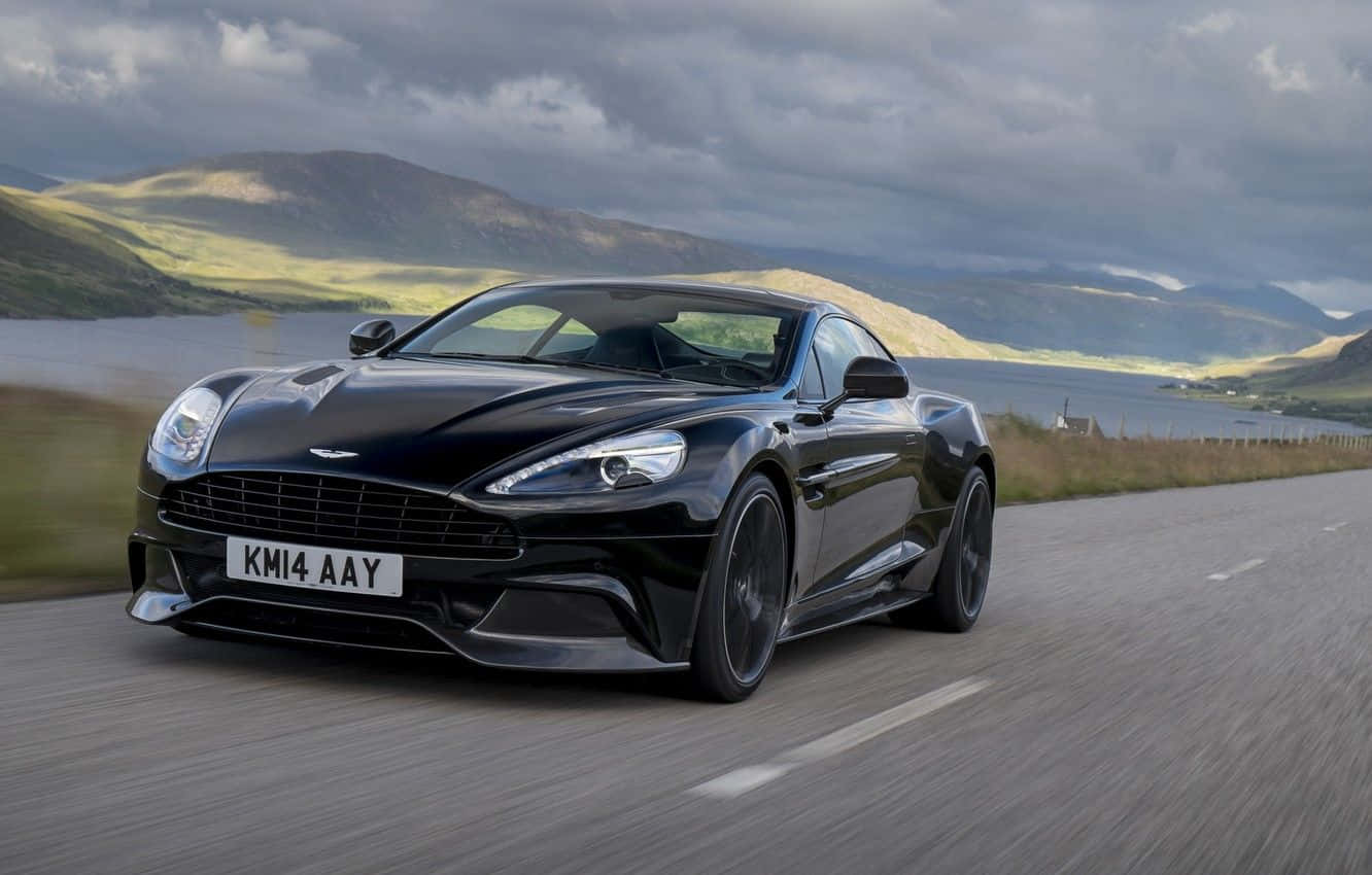 Aston Martin Vanquish - 豪華高性能跑車 壁紙 — 一架時尚的黑色Aston Martin跑車在蜿蜒的道路上奔馳，兩旁是起伏的綠色山丘和一個湖泊，天空多雲。