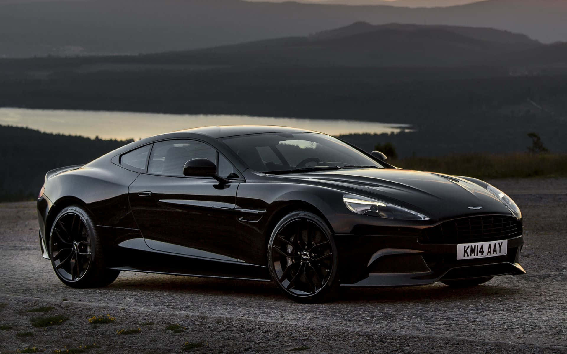 震撼嘅Aston Martin Vanquish行駛中 壁紙 — 一架時尚的黑色Aston Martin跑車在黃昏時停泊在碎石觀景點，背景有湖泊和山丘。