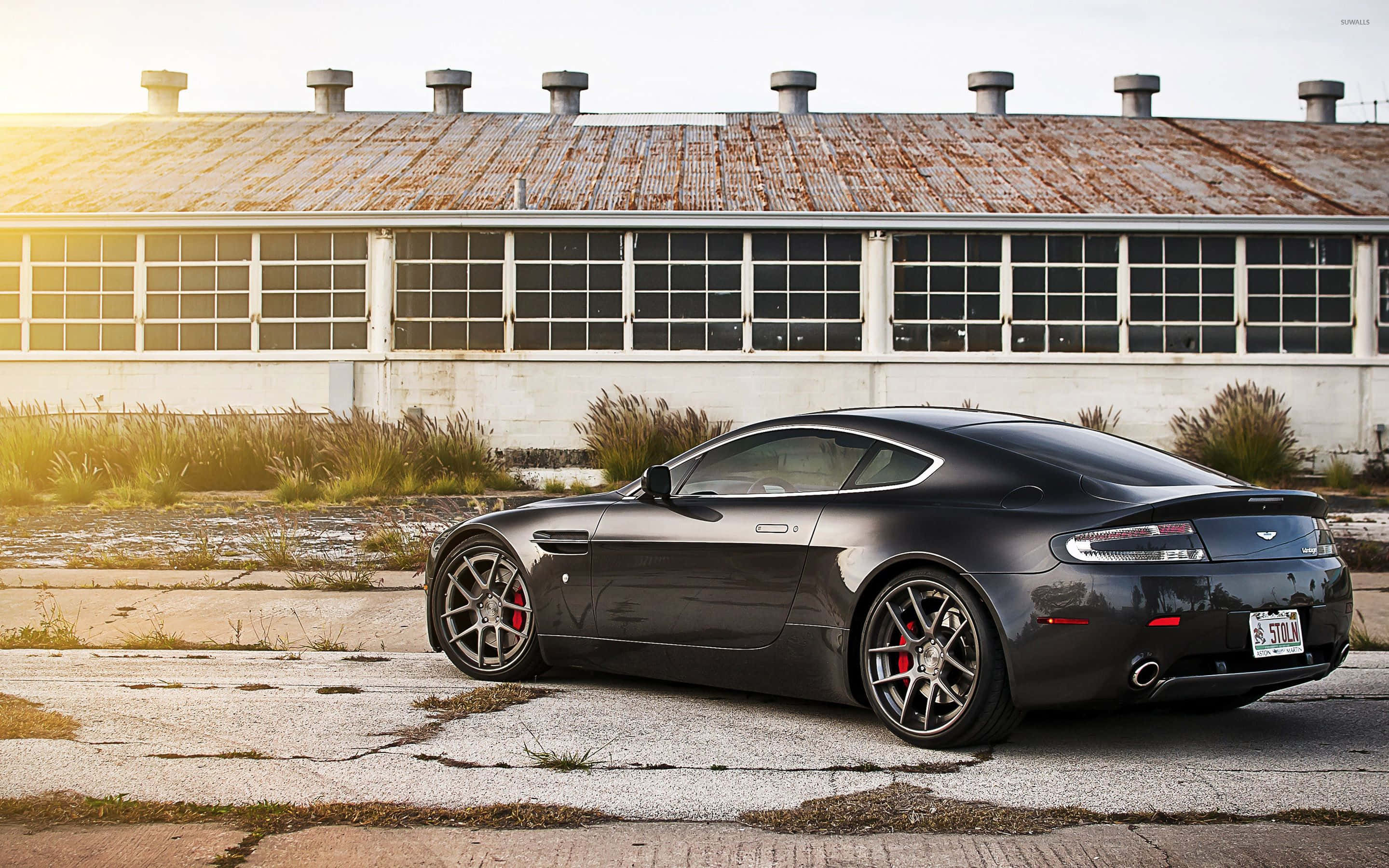 Aston Martin Vanquish喺風景靚路上馳騁 壁紙 — 一輛時尚的黑色Aston Martin跑車停在金黃時分、生鏽工業建築前破裂的路面上。