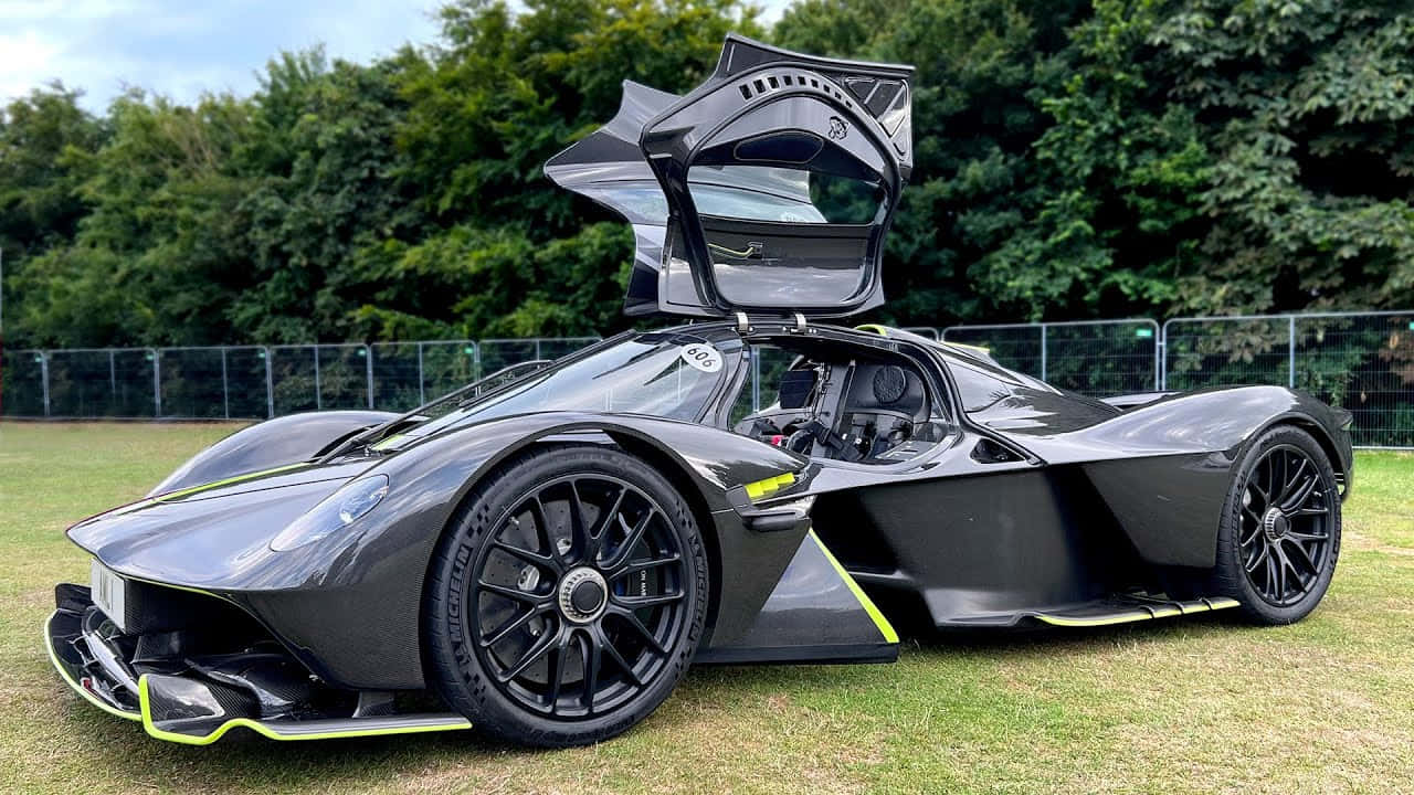 Aston Martin 出現在豪華車展 壁紙 — 一輛時尚的黑色超級跑車，帶有發光的黃色點綴和戲劇性的向上開啟車門，停在草地上，天空多雲。