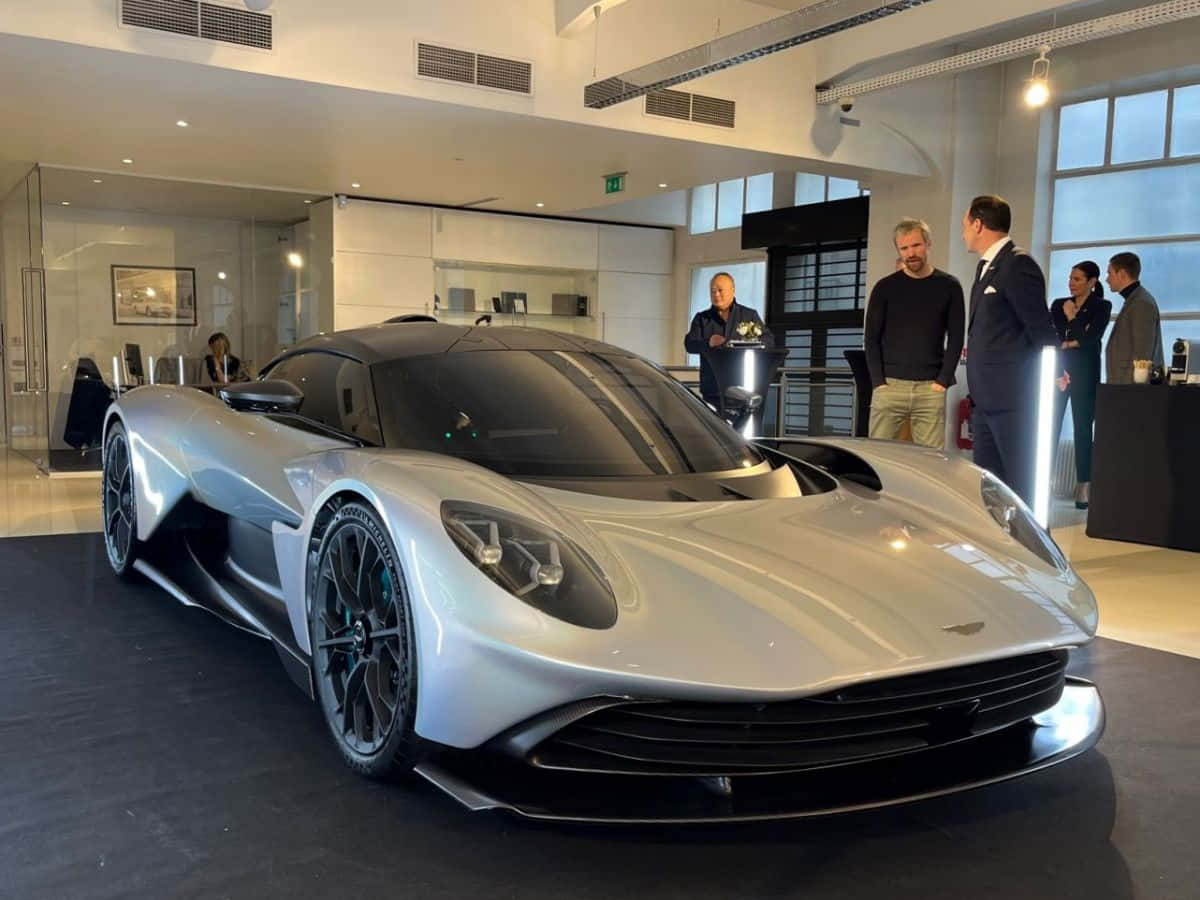 Aston Martin 的創新奢華 壁紙 — 一架流線型的銀色Aston Martin超跑在明亮燈光下閃閃發光，一群男士在現代豪華陳列室中欣賞它。