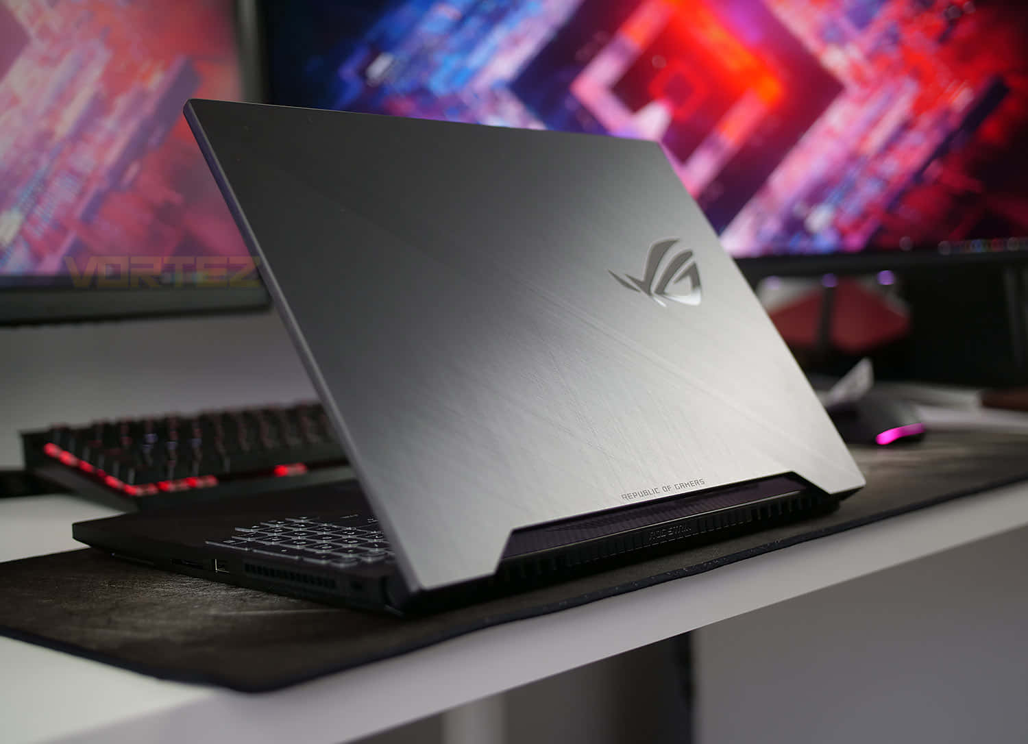 體驗 ASUS Republic of Gamers 的極致遊戲效能 壁紙 — 一部時尚的銀色遊戲筆記本電腦打開放在桌上，背景有發光的紅色鍵盤和鮮豔的RGB顯示器。