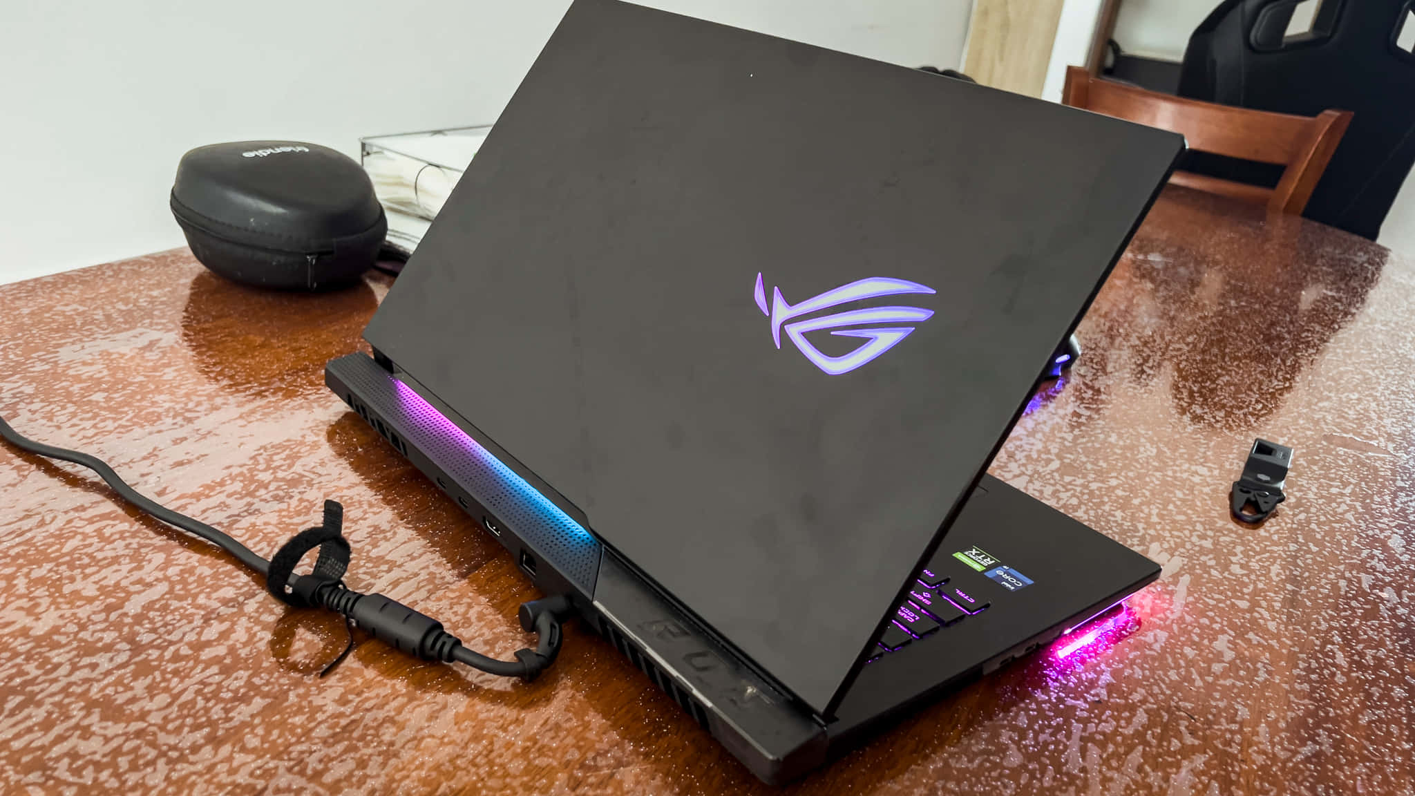體驗強勁升級的遊戲效能，選擇 ASUS ROG 壁紙 — 一部黑色 ROG 電競手提電腦，帶有發光的紫色和藍色 RGB 燈光，打開放在一張有斑點的棕色桌子上，附近有耳機和保護套。