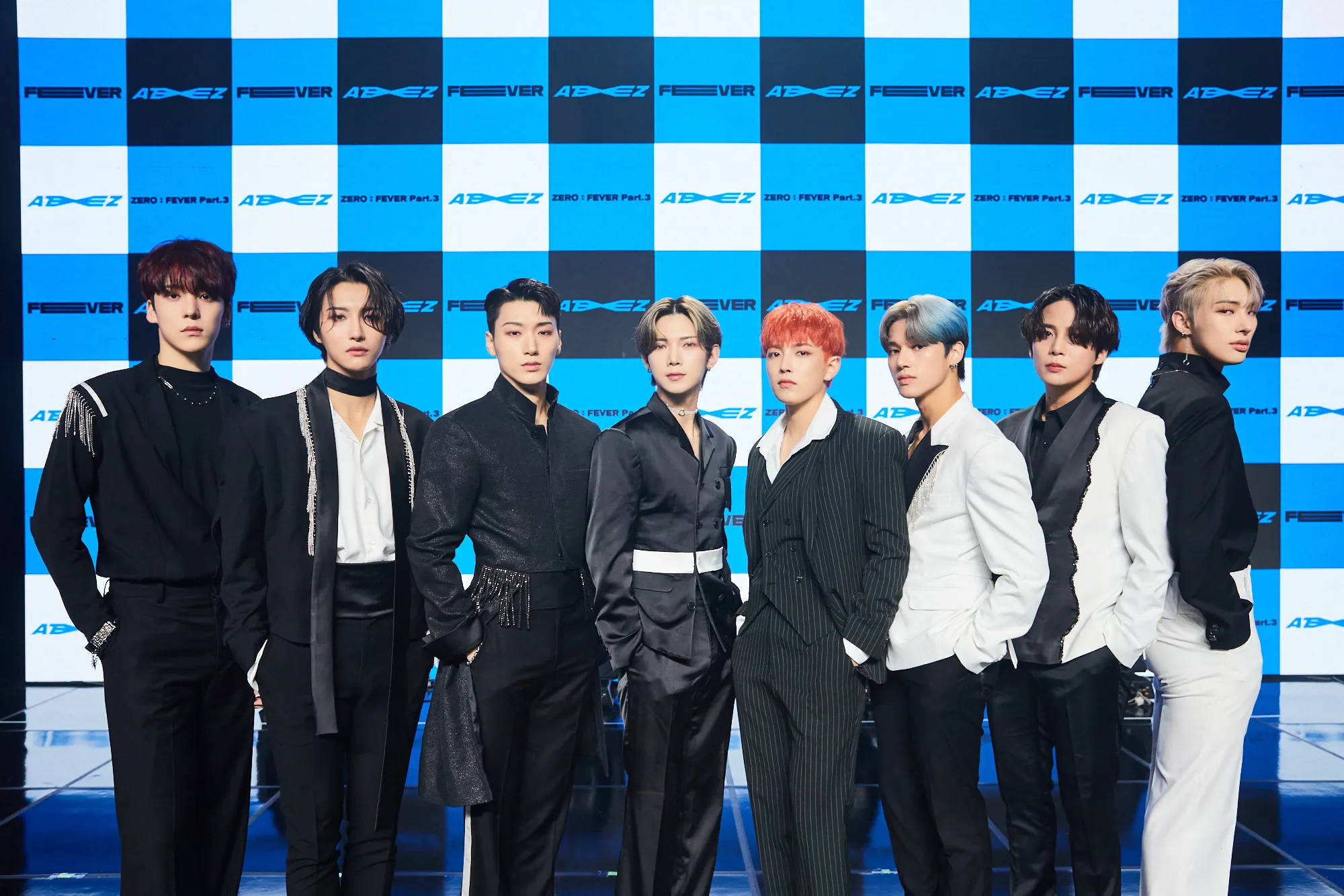 七位K-pop偶像穿著時尚的黑白西裝，在藍色格仔舞台背景上擺姿勢。