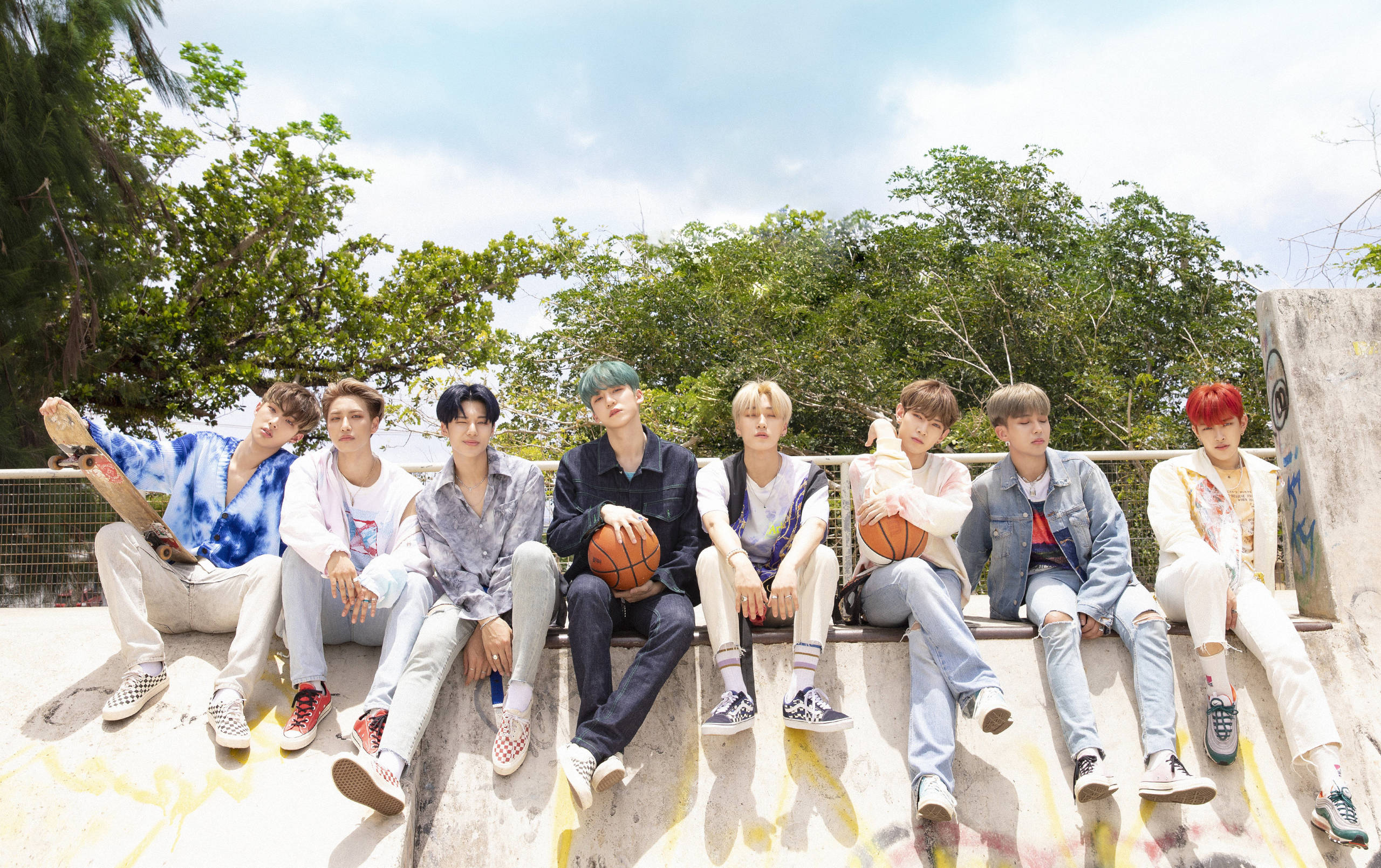 Ateez坐於藍天桌面 壁紙 — 七個年輕男子穿著休閒街頭服飾，放鬆地坐在一個被塗鴉覆蓋的混凝土邊緣上，背景是明亮的藍天和茂密的綠樹。