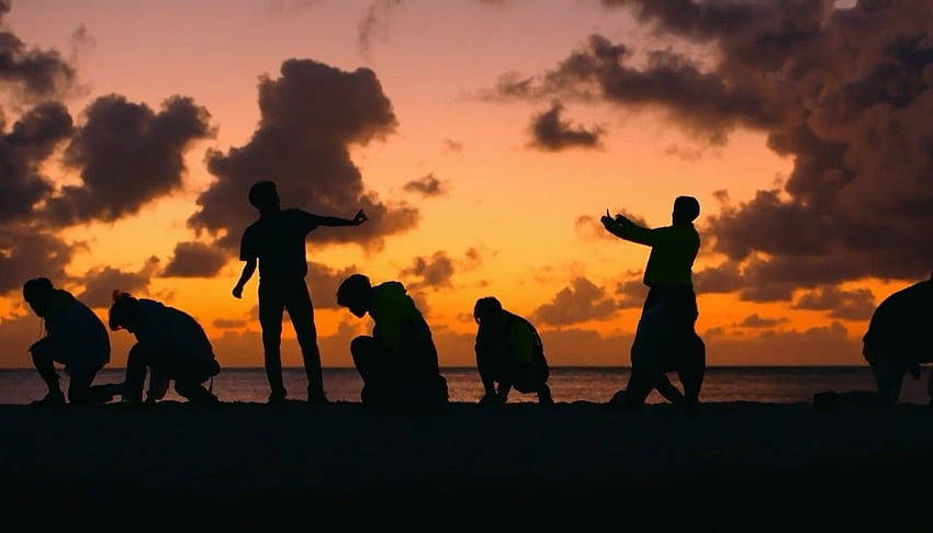 ATEEZ夕陽剪影桌面 壁紙 — 日落時分，沙灘上剪影人物擺出動態姿勢，背景是火橙色和紫色的雲彩。