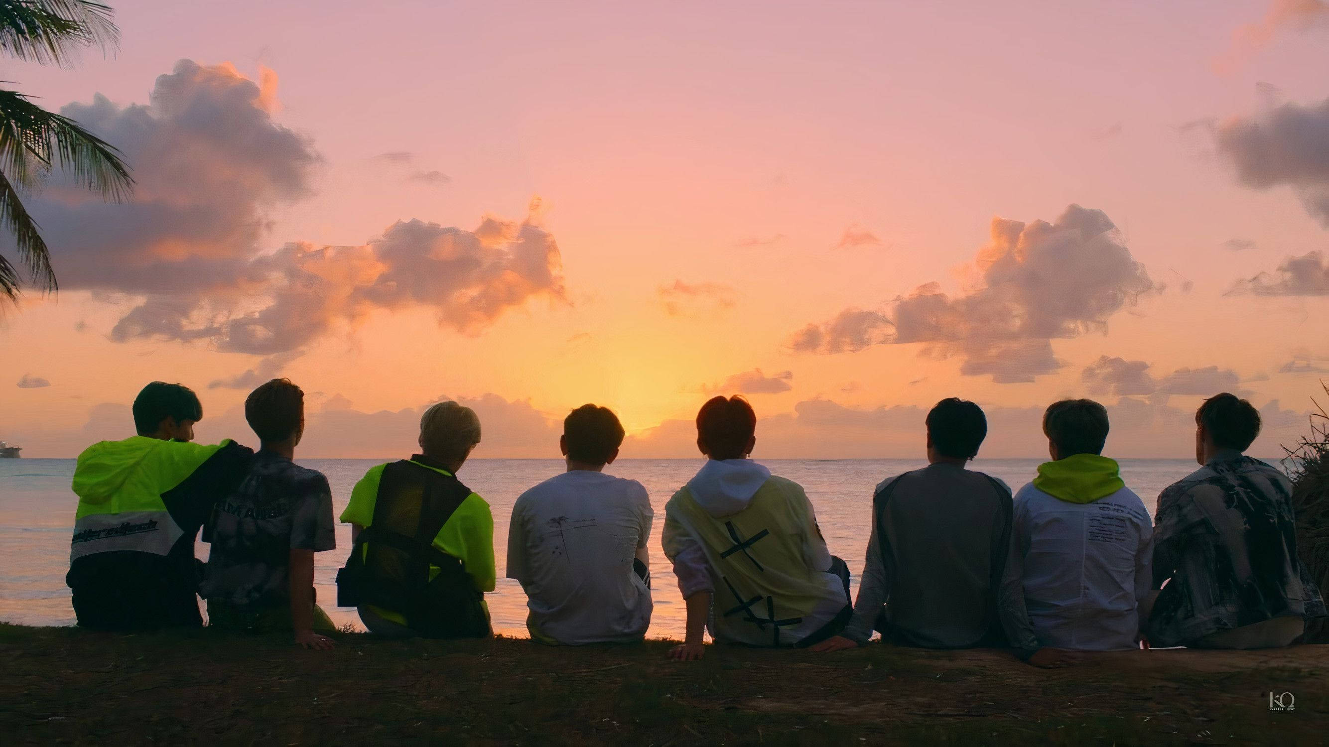 Ateez夕陽背影桌面 壁紙 — 七個年輕男子以剪影坐在沙灘上，觀賞海洋上充滿活力的橙粉紅色日落，左側有棕櫚葉框住。