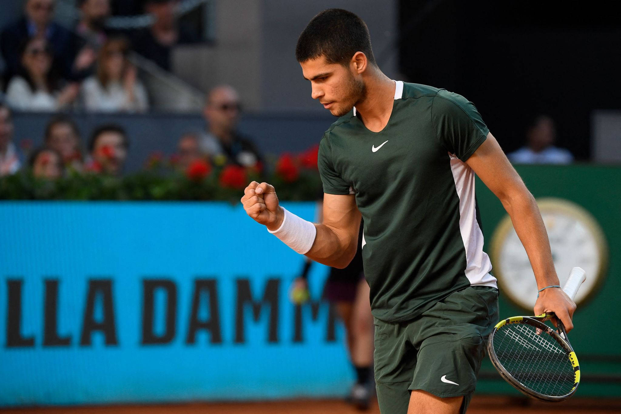 網球選手卡洛斯·阿爾卡拉斯 ATP 巡迴賽 壁紙 — 一位專注的網球選手身穿綠色裝備，在紅土場上握拳慶祝勝利，背景觀眾模糊。