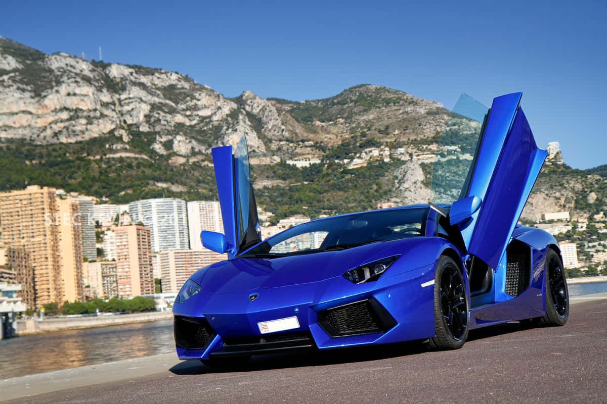 戶外藍色蘭博基尼Aventador 壁紙 — 一輛鮮藍色的林寶堅尼Aventador，剪刀門打開，停泊在沿海公路上，背景是山脈和城市建築，天空清澈湛藍。