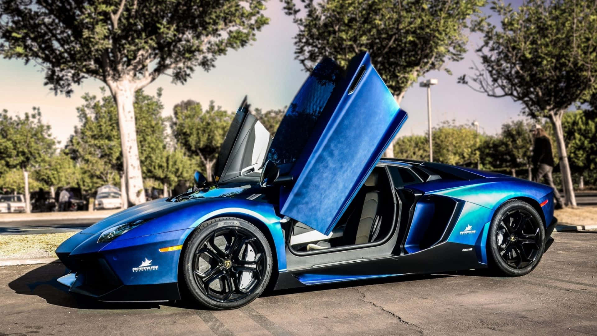 藍色蘭博基尼 Aventador 外觀酷炫 壁紙 — 一架充滿活力的藍色林寶堅尼，剪刀門打開，停泊在陽光普照、樹木成蔭的停車場。