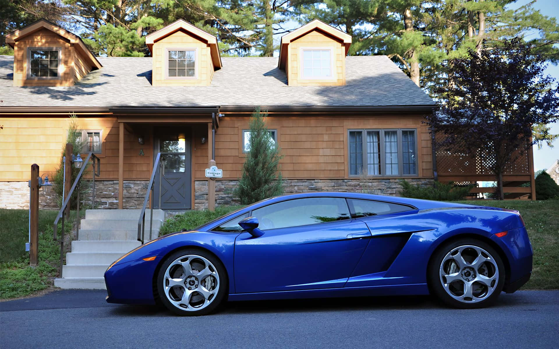 極致風格與速度——藍色蘭博基尼Aventador 壁紙 — 一架充滿活力的藍色林寶堅尼Gallardo停泊在一間帶有石頭裝飾和高大樹木的質樸木屋前的車道上。
