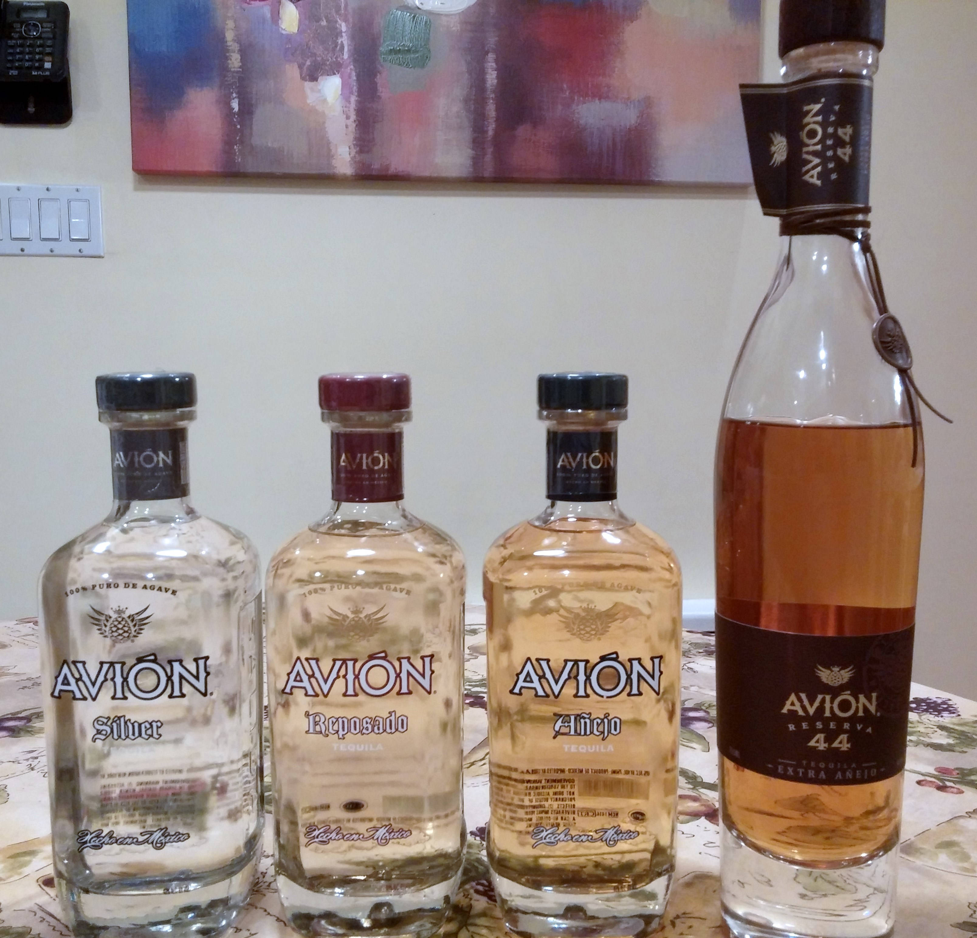 Avion 蒂奎拉酒 44 與其他系列 壁紙 — 四瓶不同年份的Avion龍舌蘭酒在溫暖的室內燈光下排列在花卉桌布上。