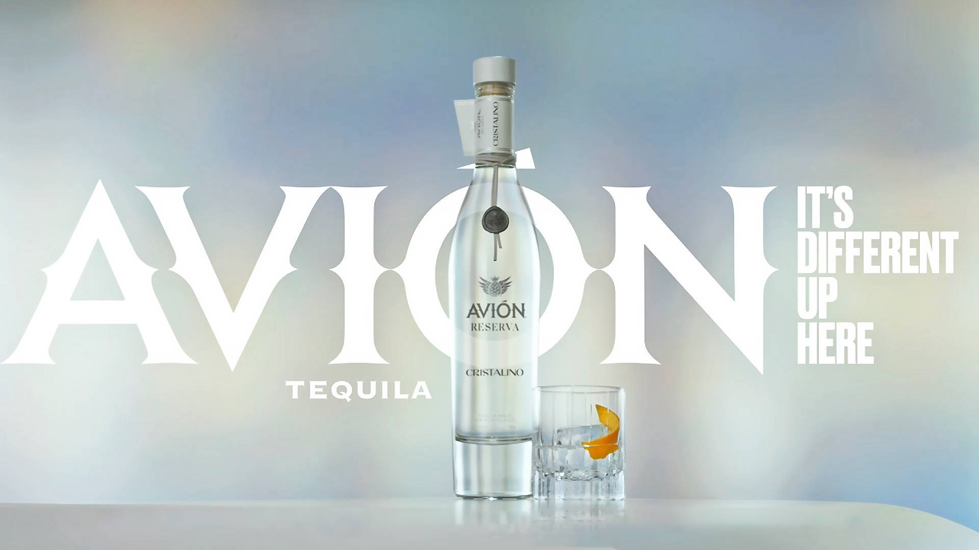 Avion 龍舌蘭酒 Cristalino 壁紙 — 一個時尚的Avion龍舌蘭酒瓶和帶有橙片的玻璃杯放在白色表面上，背景為柔和的漸變色，並帶有粗體白色字體。