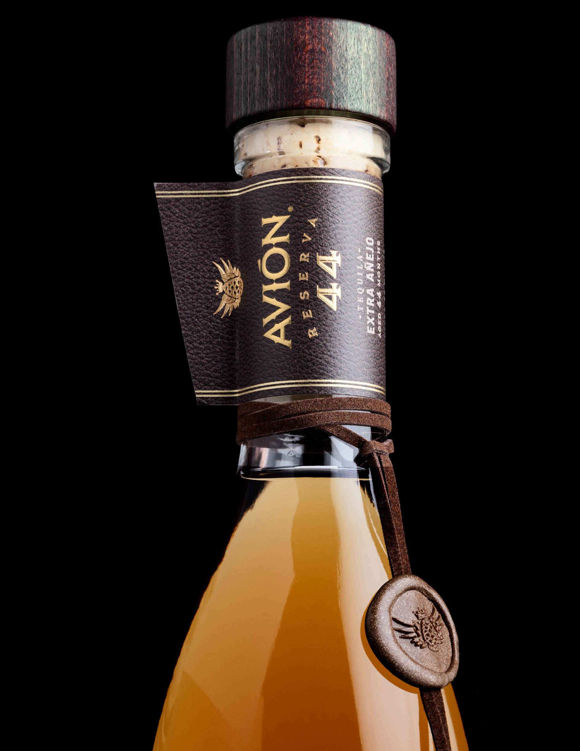 奢華 Avion 蒸餾酒靜置於樸素背景 壁紙 — Avion Reserva 44 龍舌蘭酒瓶特寫，琥珀色液體、皮革標籤和木蓋，黑色背景。