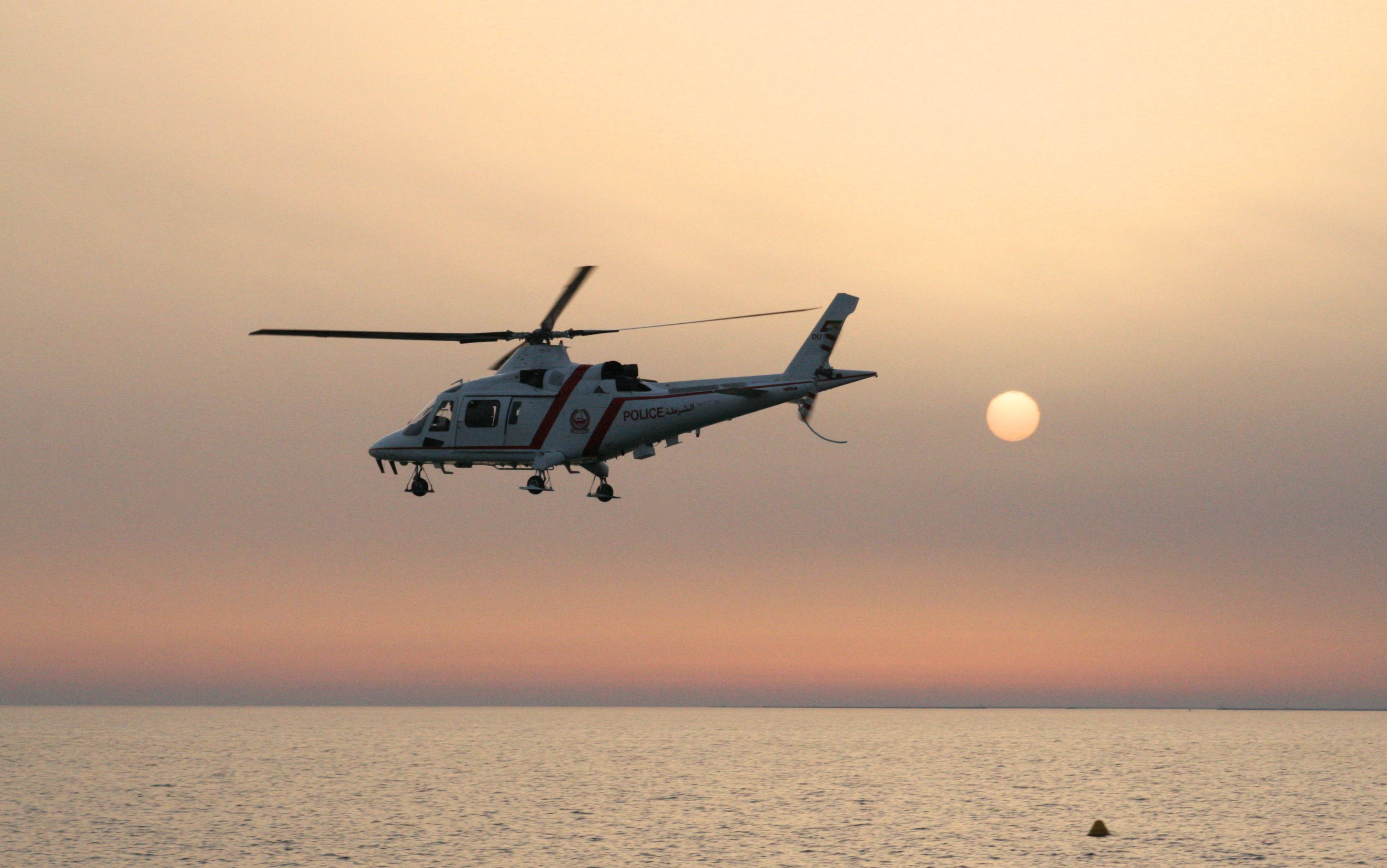 AW109直升機4K 壁紙 — 一架警用直升機在平靜的海面上盤旋，夕陽在柔和的橙粉色的天空中緩緩落下。