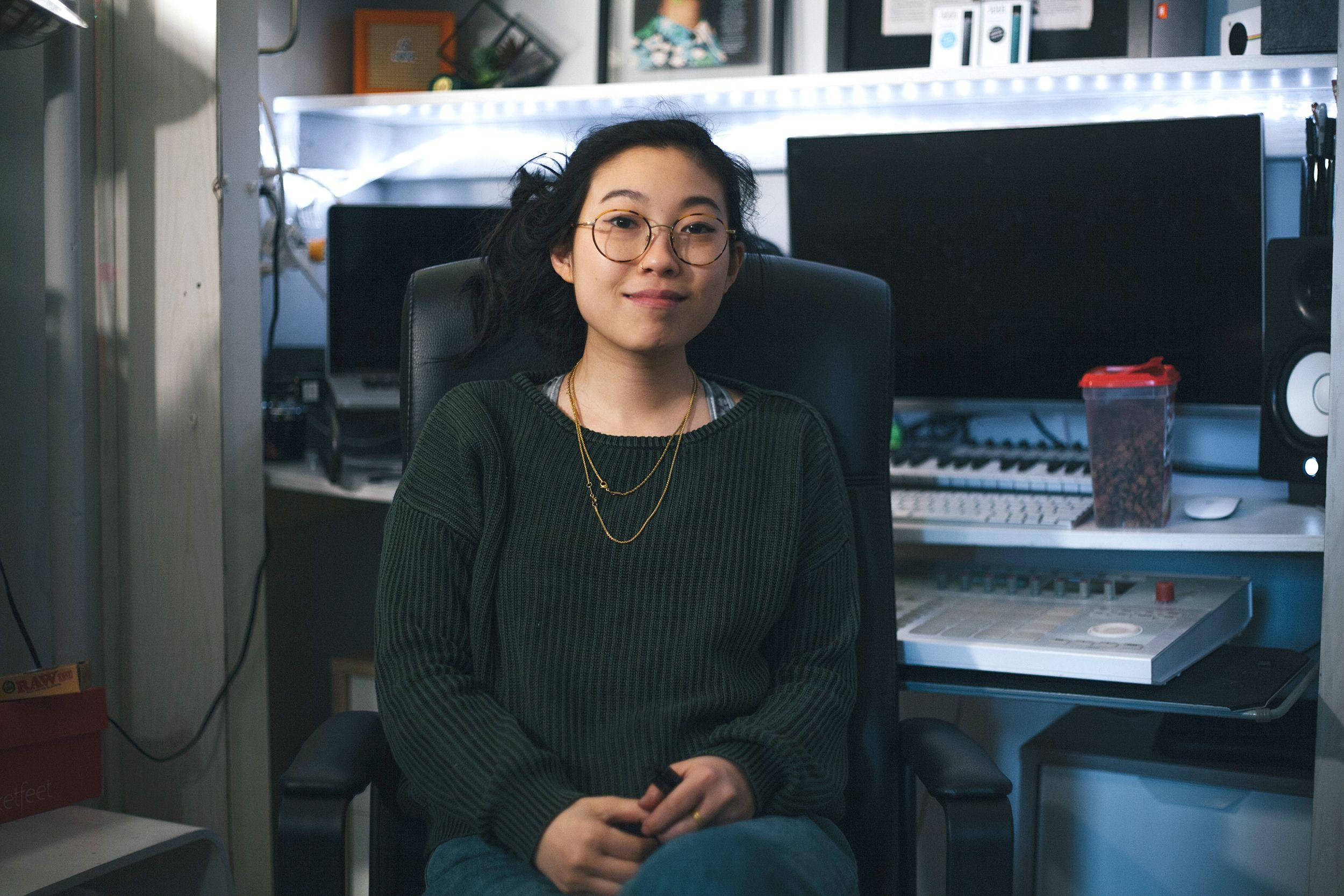 Awkwafina 在音樂工作室內 壁紙 — 一位戴眼鏡的年輕女性微笑著坐在黑色辦公椅上，身處充滿科技感的家庭工作室，沐浴在柔和的藍色LED燈光中。