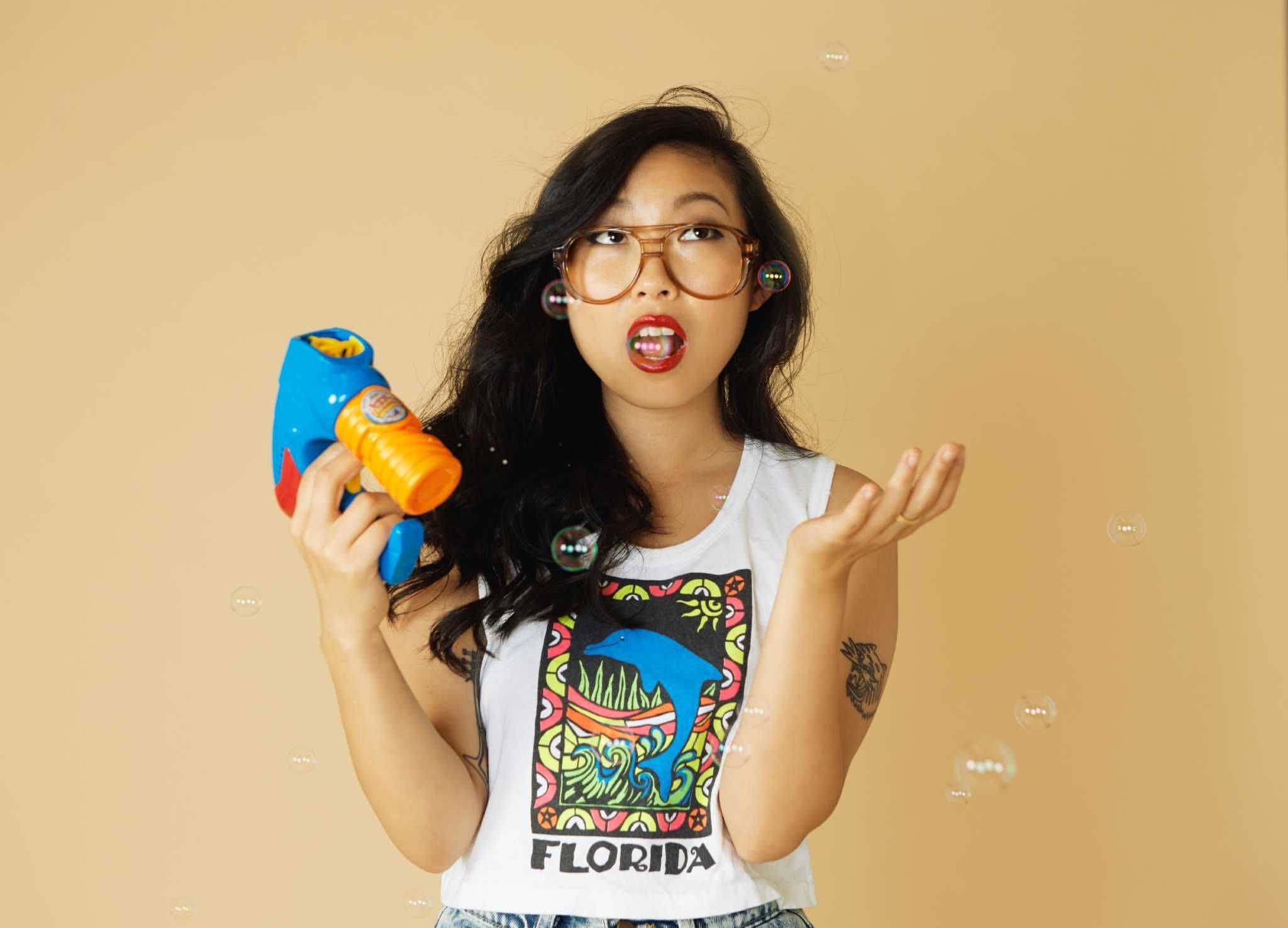 Awkwafina 手持泡泡槍 壁紙 — 一位戴眼鏡、身穿佛羅里達背心的年輕女性，在米色背景前俏皮地吹泡泡。