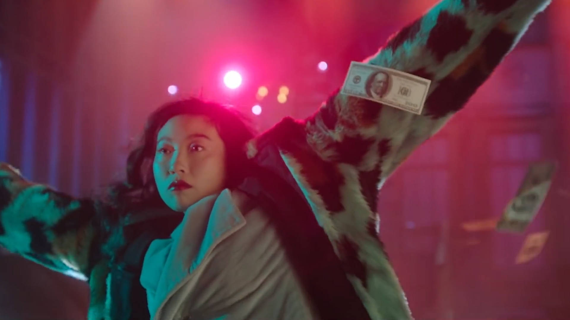 Awkwafina 穿毛皮夾克 壁紙 — 一個人穿著豹紋外套，在充滿活力的紅色和藍色舞台燈光下將錢拋向空中。