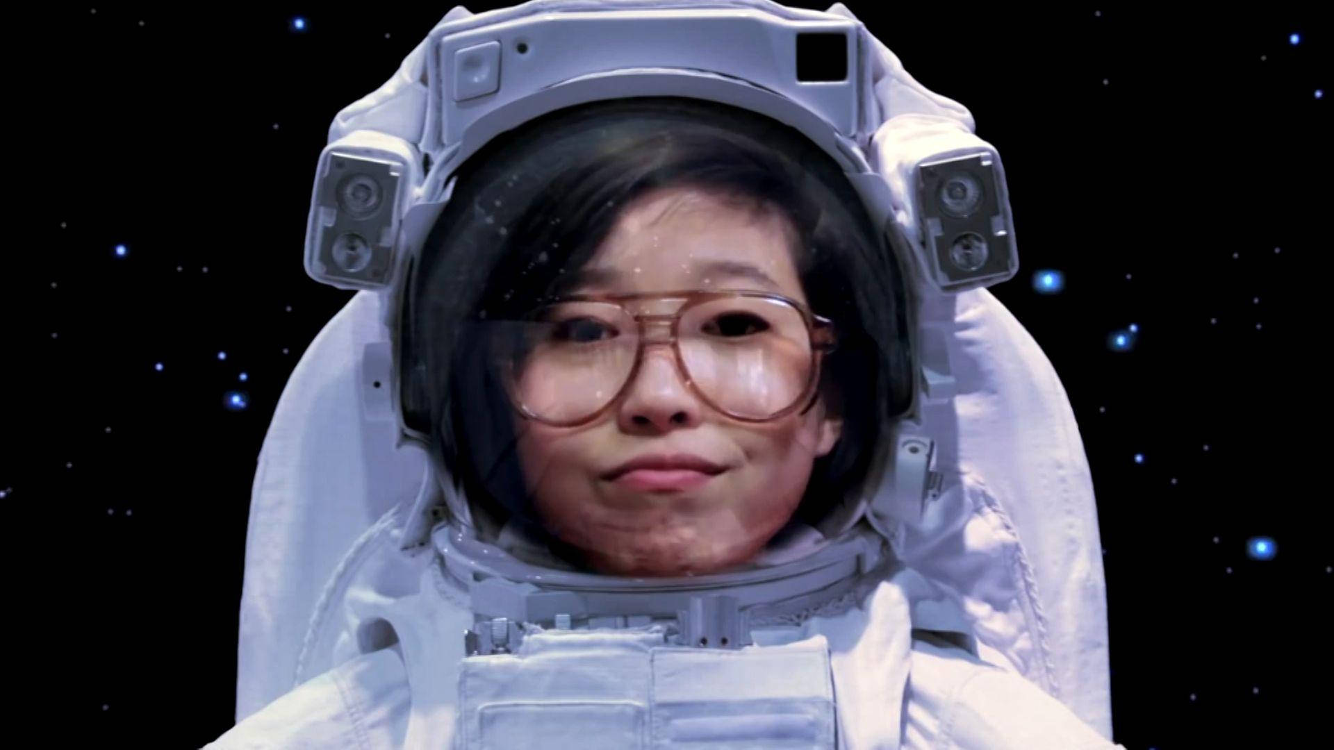 Awkwafina 穿白色太空服 壁紙 — 一位戴眼鏡的年輕女子身穿白色太空服，在繁星點點的黑色太空背景下平靜地凝視著鏡頭。