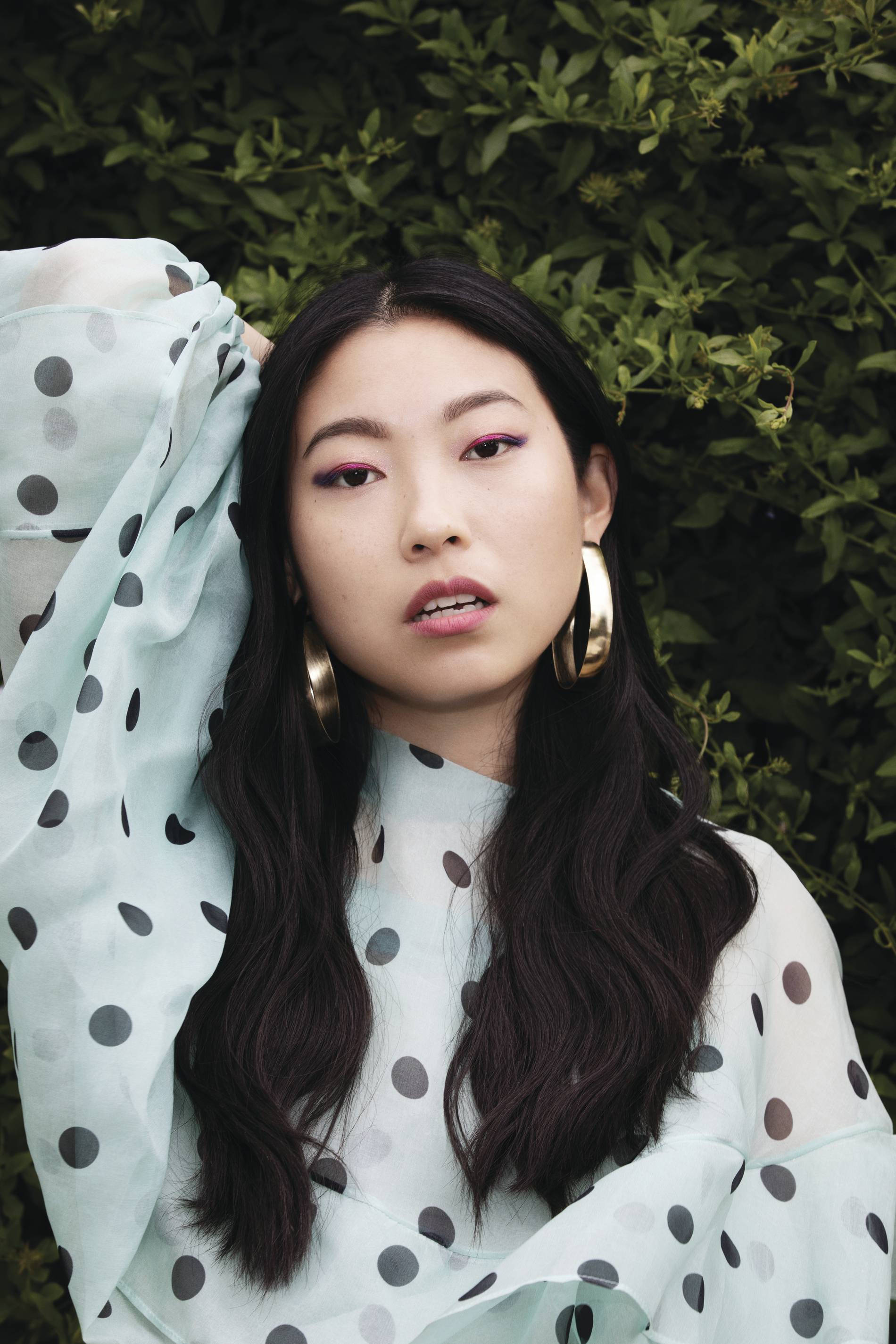 Awkwafina 穿白色波點連身裙 壁紙 — 一位深色頭髮、妝容大膽的年輕女性，在茂密的綠色樹籬前擺姿勢，身穿淺藍色圓點襯衫和金色耳環。