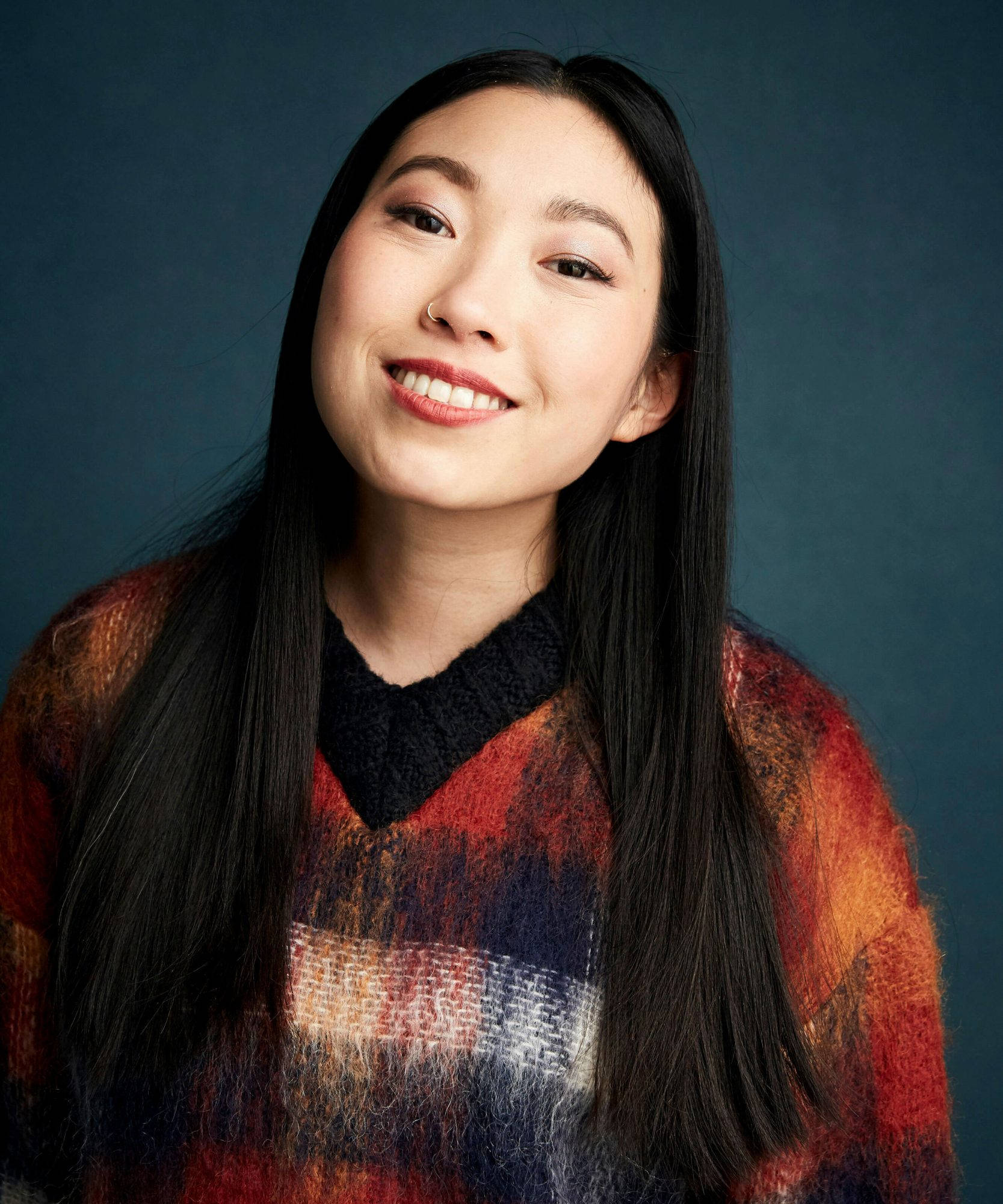Awkwafina 紅橙格紋毛衣 壁紙 — 一位微笑的年輕女性，長長的黑髮，穿著色彩繽紛的質感毛衣，背景是深藍色的攝影棚。