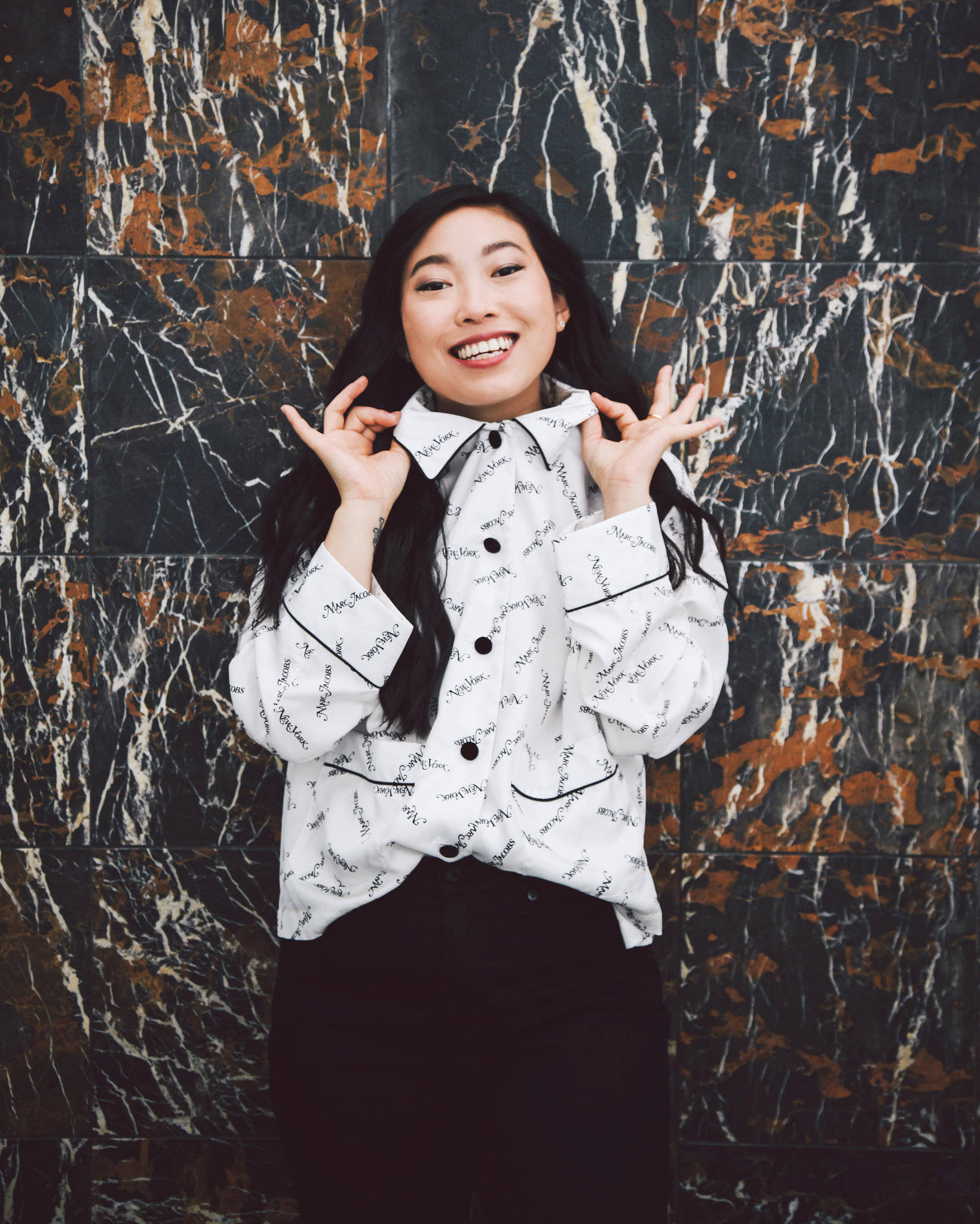 Awkwafina 超大POLO衫 壁紙 — 一位長黑髮的快樂女性，在引人注目的黑白金大理石牆前燦爛微笑擺姿勢。