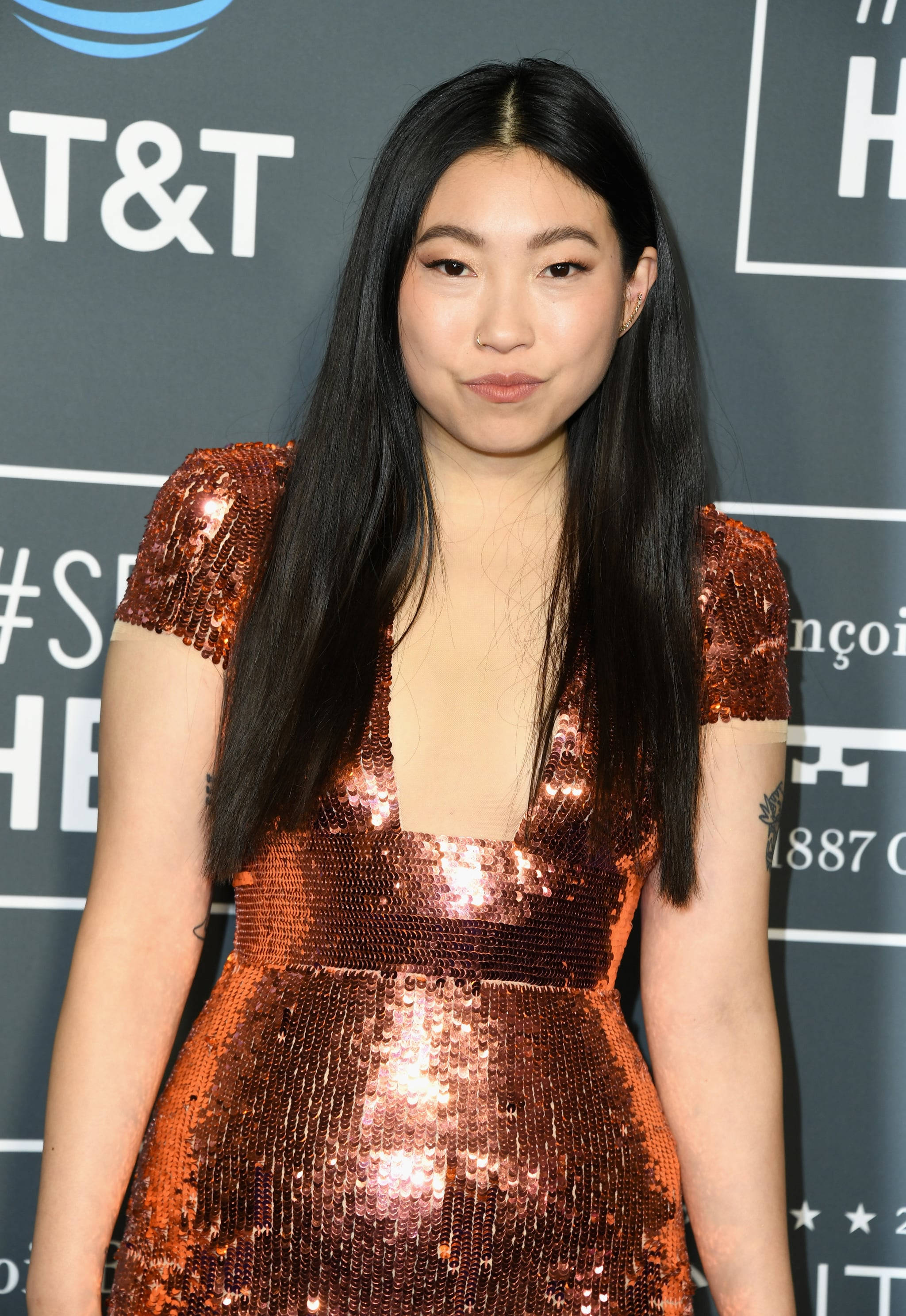 Awkwafina 金屬銅色亮片禮服 壁紙 — 一位長黑髮的女性在紅地毯活動中穿著閃閃發光的銅色亮片連衣裙，背景帶有品牌標誌。
