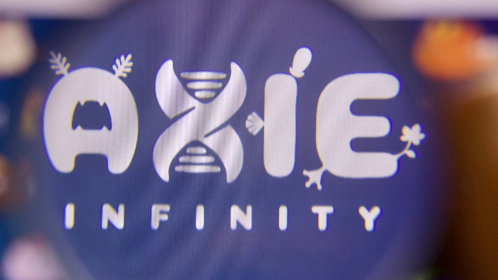 Axie Infinity 名稱標誌特寫 壁紙 — Axie Infinity 標誌特寫，在深藍色屏幕上發出白色光芒，周圍有柔和模糊的燈光。