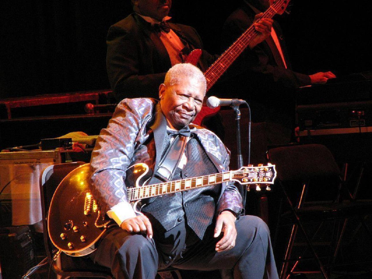 B.B. King 在多倫多Roy Thompson Hall演出 壁紙 — 一位微笑的老年音樂家穿著圖案西裝，在燈光昏暗的舞台上彈奏金色電結他，身後有一位低音結他手。
