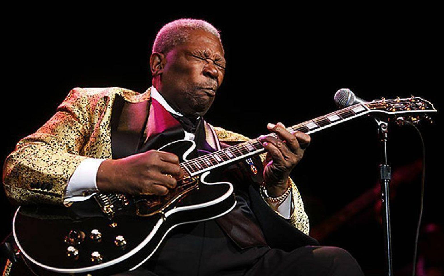 B.B. King 演奏吉他獨奏 壁紙 — B.B. King 在戲劇性的舞台燈光下，穿著閃閃發光的金色外套，熱情地彈奏他的黑色電結他。