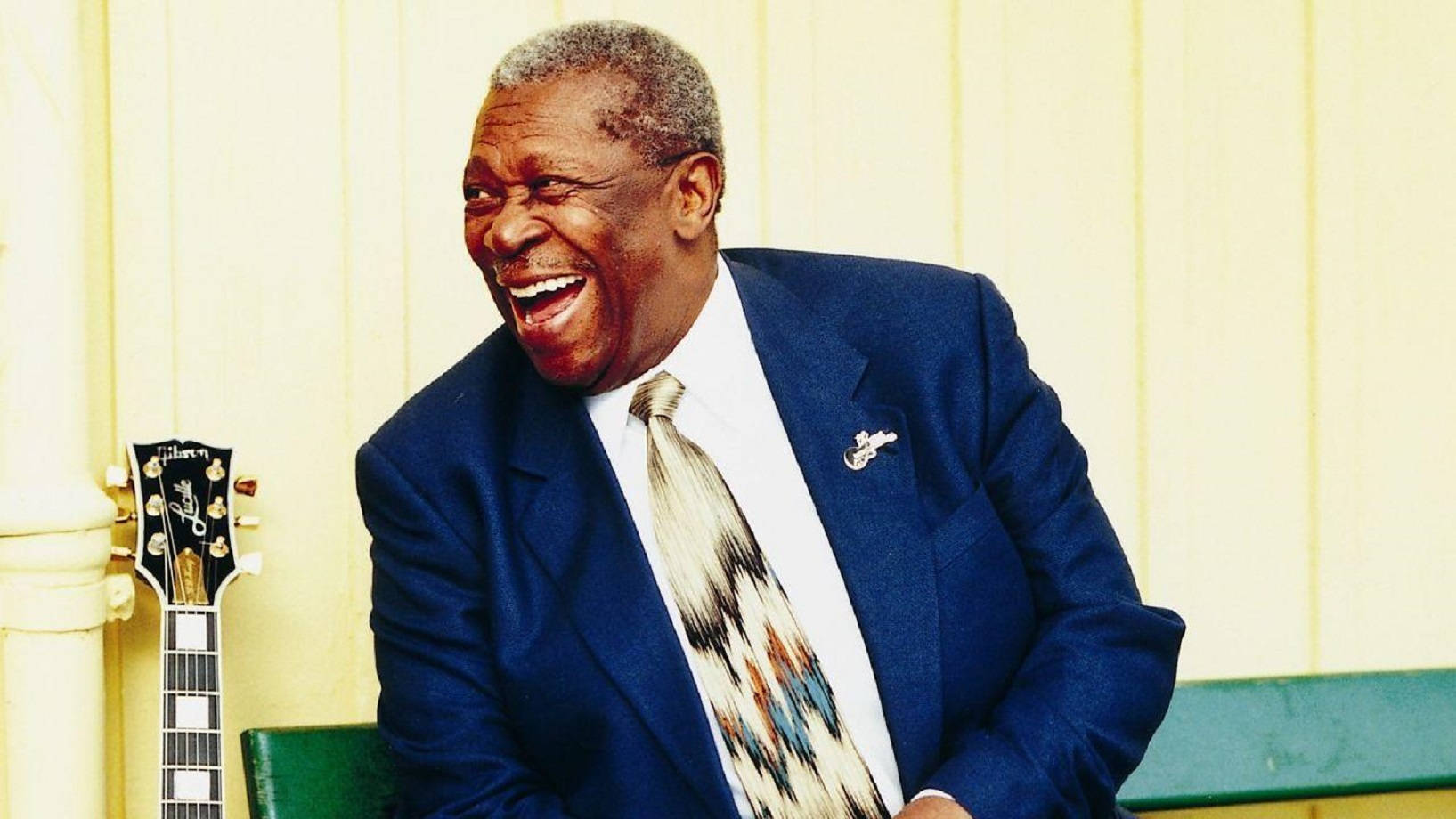 B.B. King 靠近他的吉他Lucille 壁紙 — 一位身穿藍色西裝的年長黑人男子在淡黃色牆壁旁，對著結他開懷大笑。