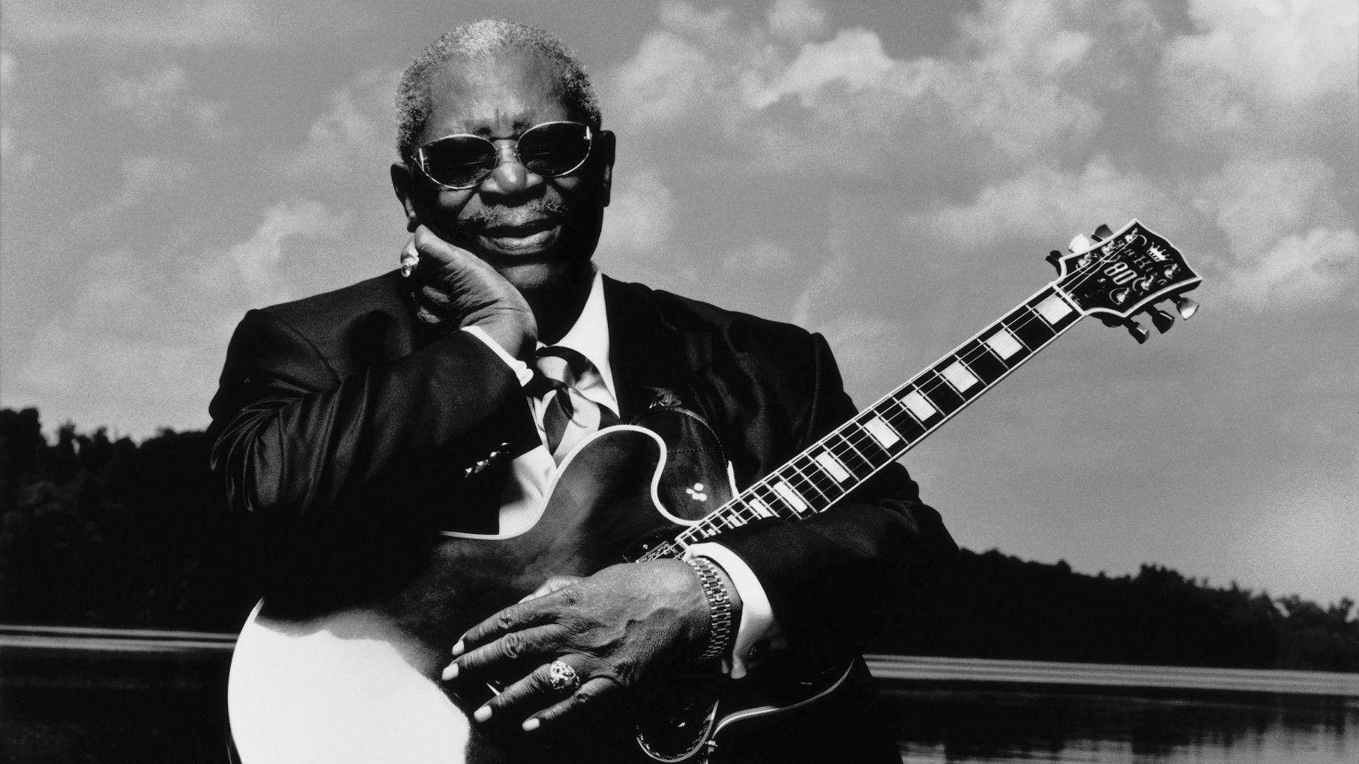 B.B. King宣傳攝影 壁紙 — 一位傳奇爵士結他手，身穿筆挺西裝和太陽眼鏡，在陰天和水景背景下手持電結他。