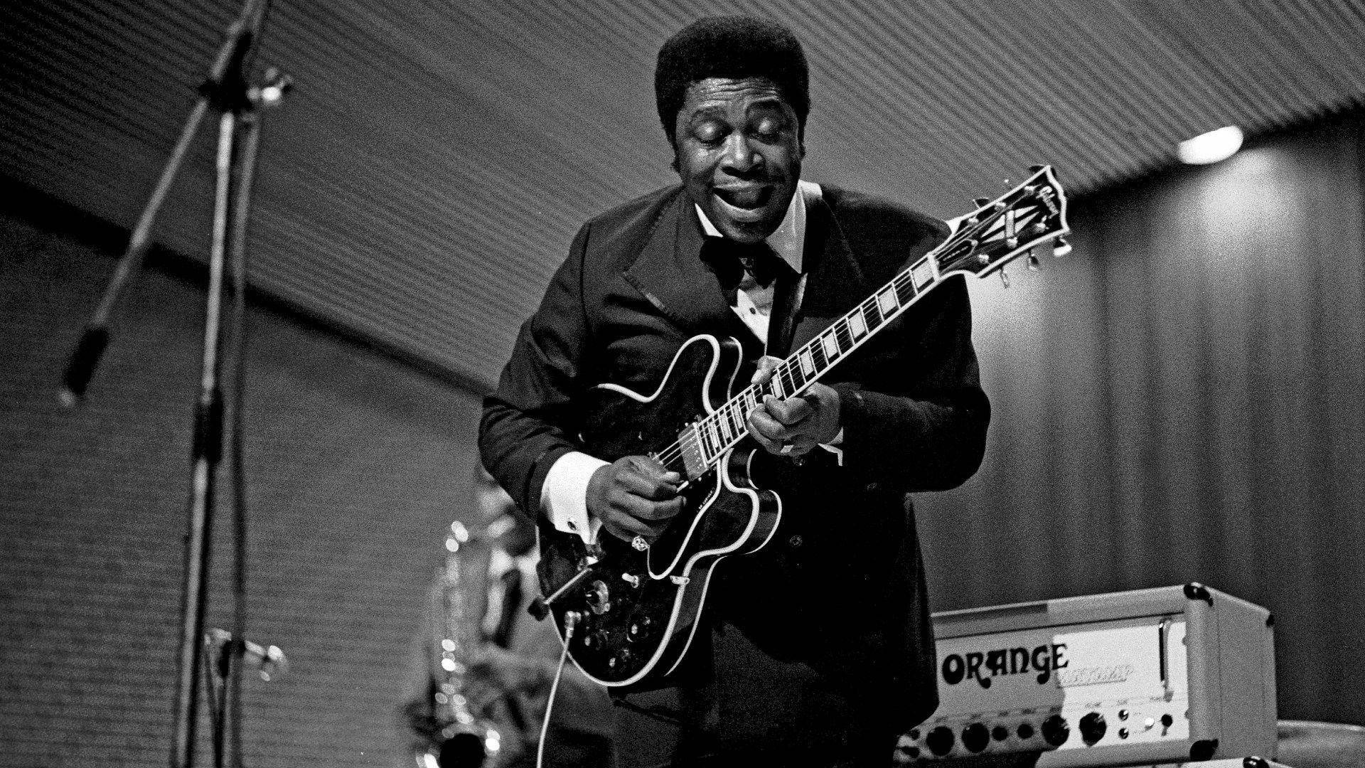 B.B. King於德國演出 壁紙 — 一位身穿西裝的快樂音樂家在舞台上熱情地彈奏電結他，身後有一個橙色擴音器。