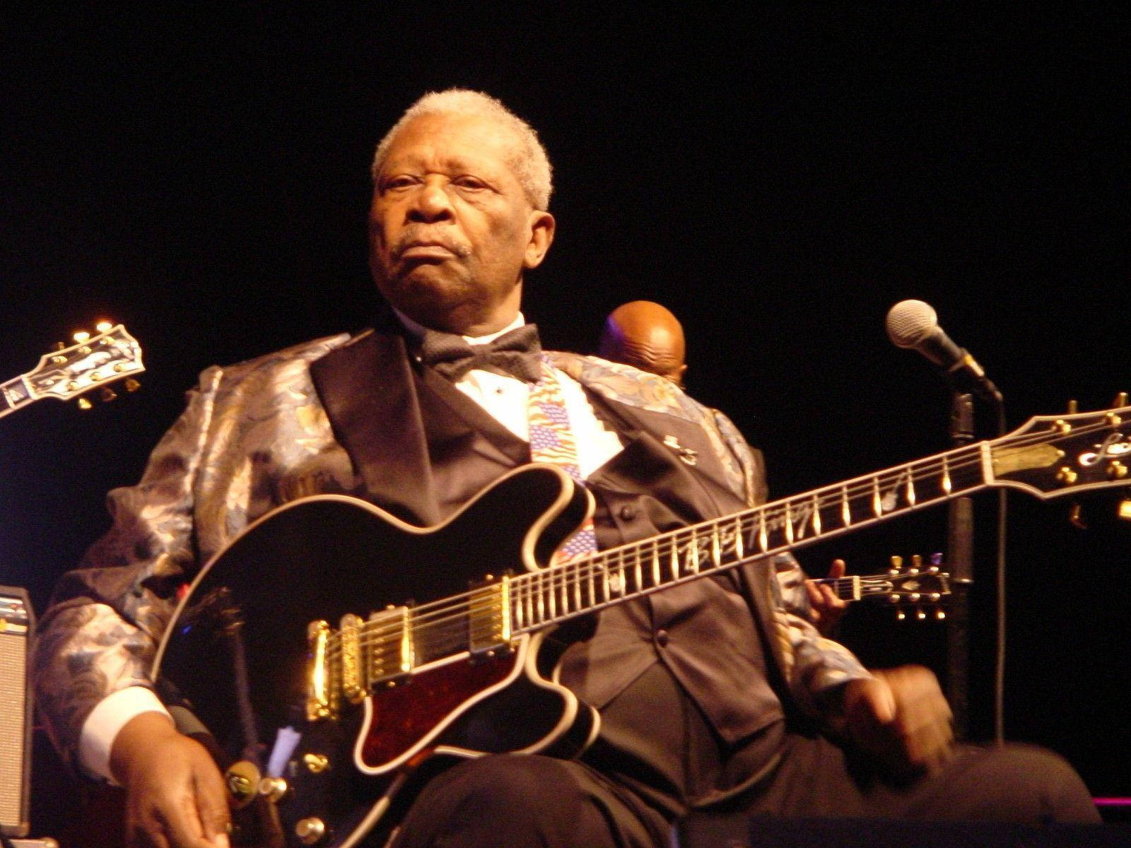 B.B. King於麥迪遜廣場花園演出 壁紙 — 一位年長的黑人男性穿著閃亮的西裝，在舞台燈光下彈奏黑色電結他，附近有一個麥克風。