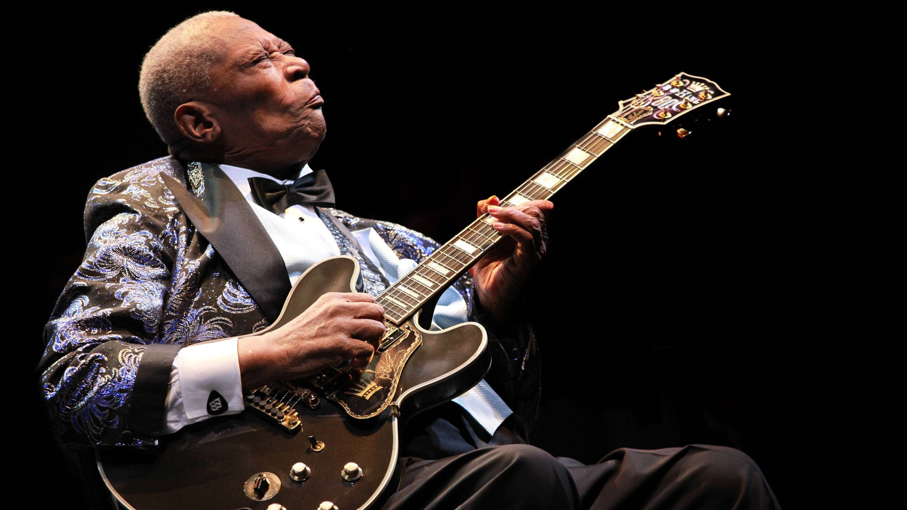 B.B. King藍調音樂節 壁紙 — 一位身穿圖案燕尾服的老年爵士結他手，在黑色背景下熱情地彈奏一把旭日紋電結他。