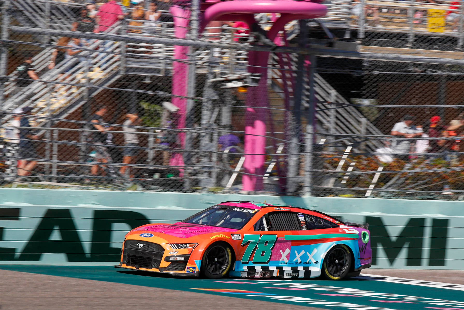 B.J. McLeod駕駛粉紅NASCAR賽車 壁紙 — 一輛充滿活力的賽車，帶有醒目的粉紅色、橙色和藍色圖案，在賽道上飛馳，觀眾在圍欄後面。