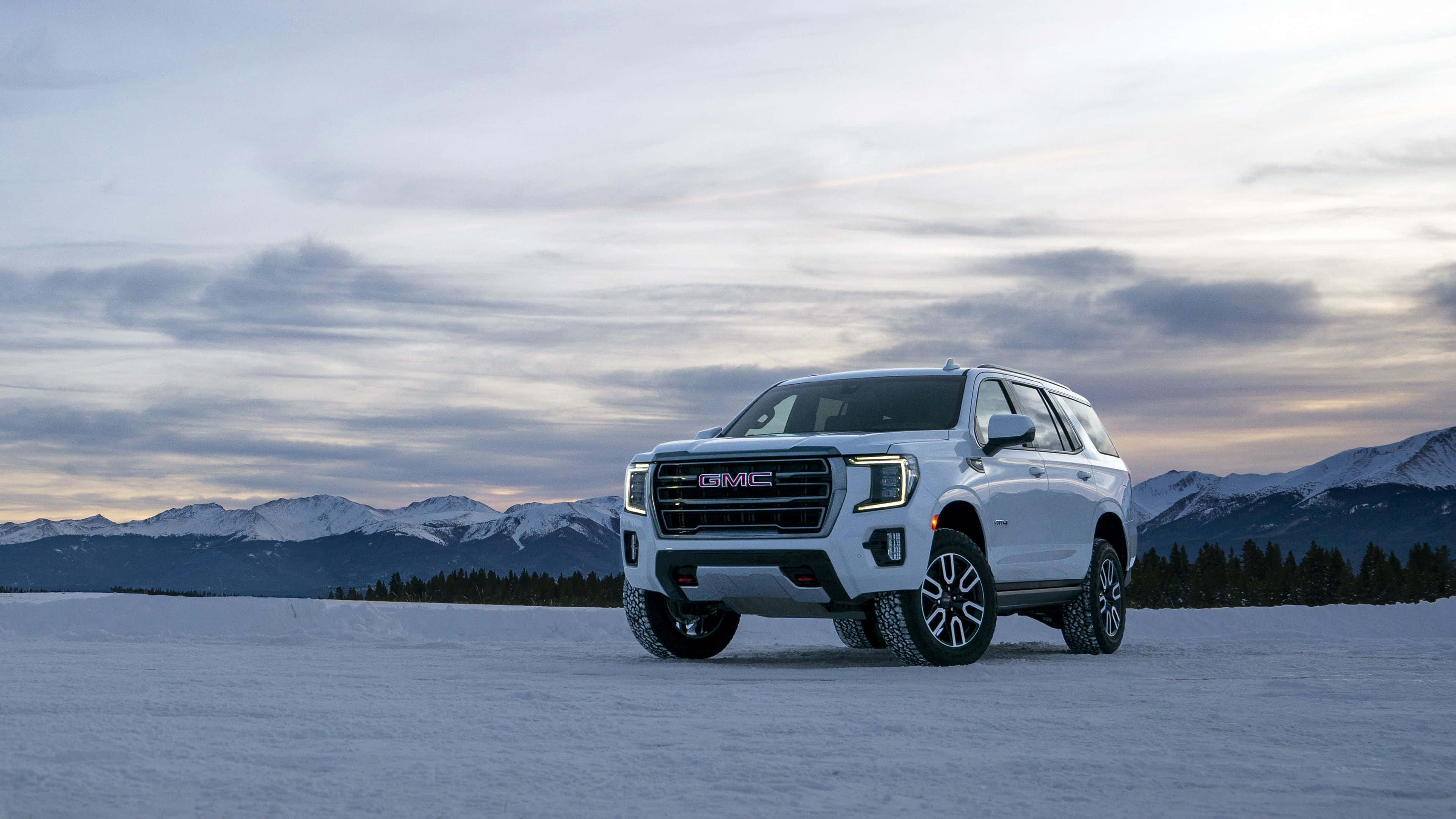Gmc Yukon 桌布 壁紙 — 一輛白色GMC SUV矗立在廣闊的雪原上，背景是積雪的山脈，天空呈現柔和的暮色。