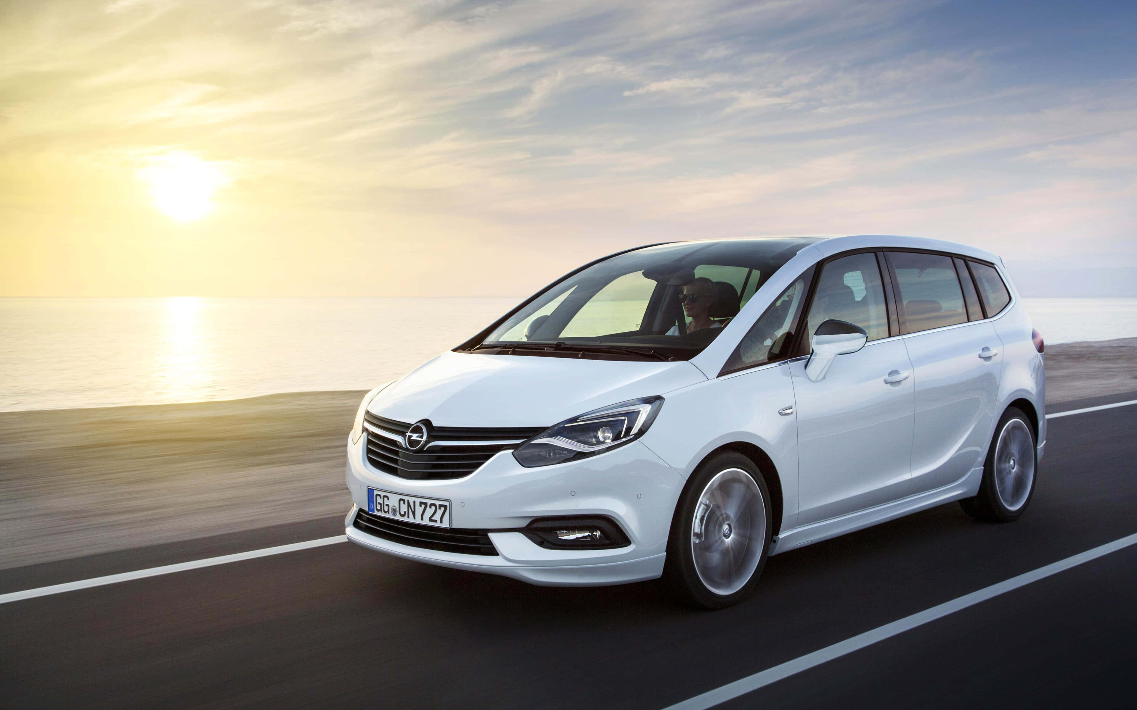 Opel Zafira 桌布 壁紙 — 一輛時尚的白色歐寶Zafira在日落時分行駛在沿海公路上，金色的光線反射在海面上。