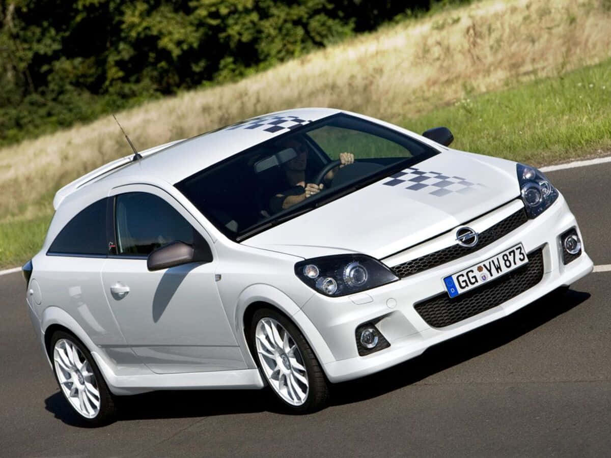 Opel Astra 桌布 壁紙 — 一輛白色歐寶雅特GTC跑車，帶有格子賽車貼紙，在明亮的陽光下沿著一條彎曲的道路疾馳，道路兩旁是綠草和樹木。