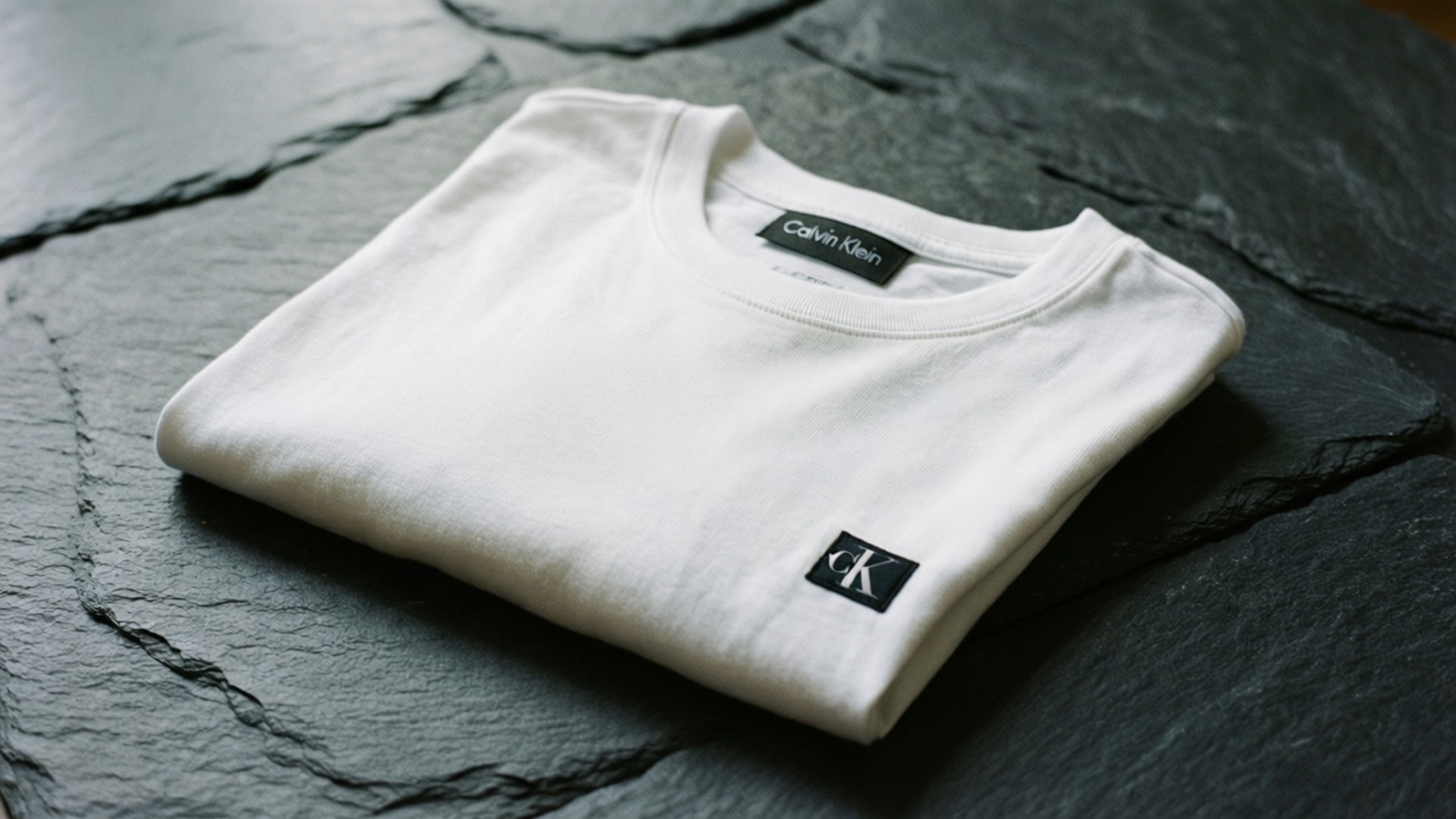 Folded Tee 壁紙 — 一件純白 Calvin Klein T 恤整齊摺疊在深色紋理石板表面上。