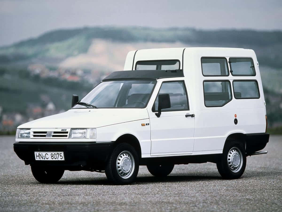Fiat Fiorino 桌布 壁紙 — 一輛白色Fiat露營車停在碎石上，背景是起伏的綠色山丘和遠處的房屋，天空多雲。