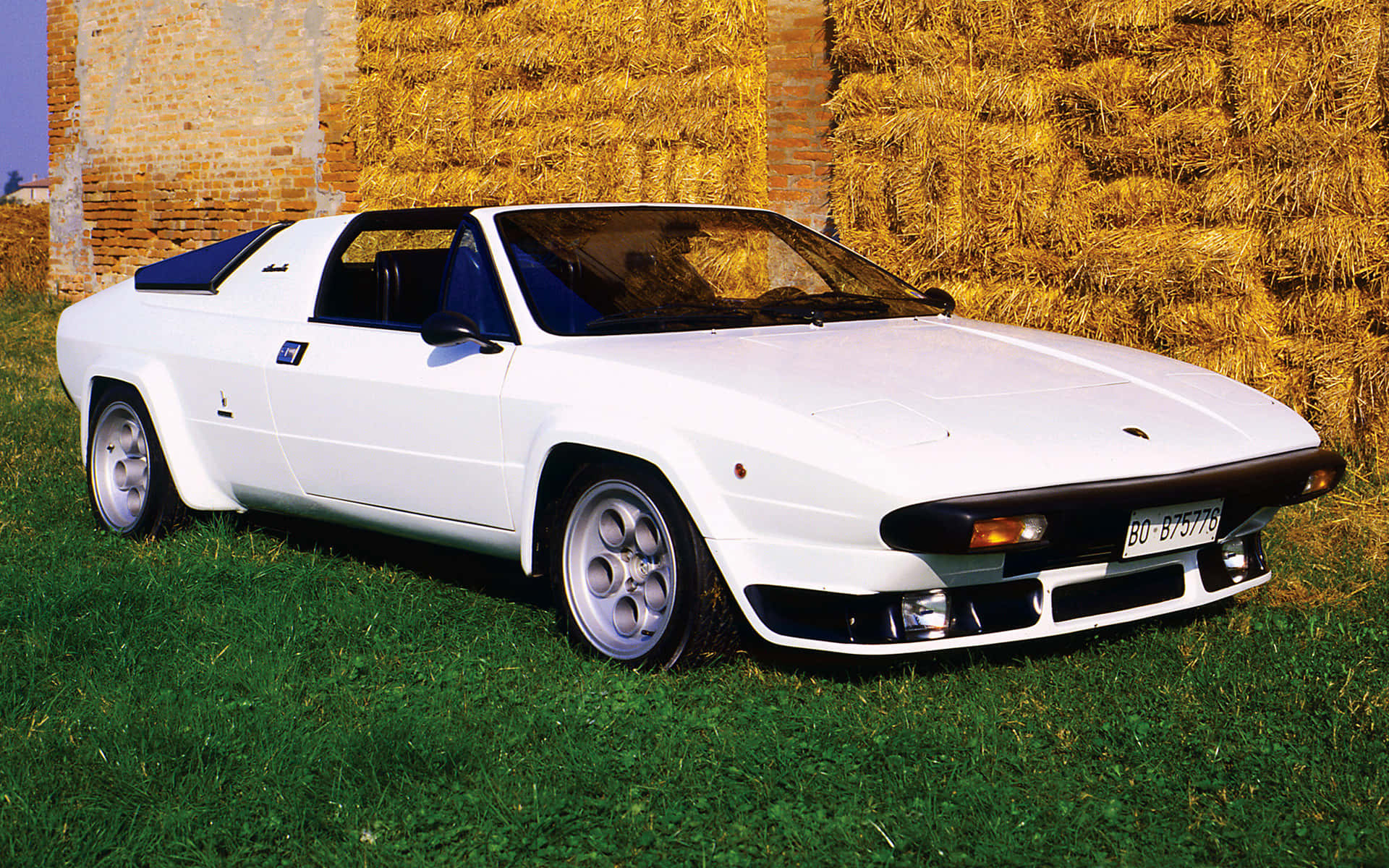 Lamborghini Silhouette 桌布 壁紙 — 一輛純白色的林寶堅尼 Jalpa 跑車停放在翠綠的草地上，旁邊是質樸的磚牆和金黃色的乾草捆。