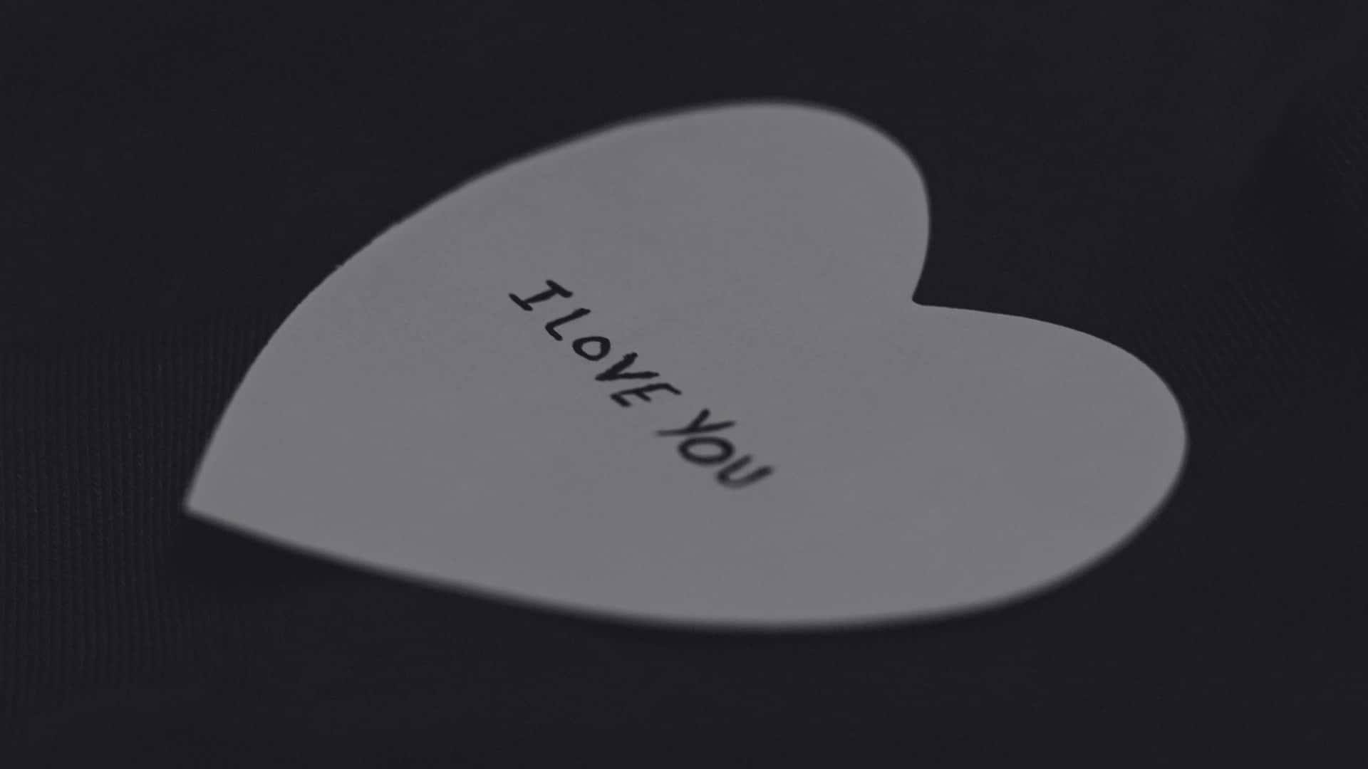  壁紙 — 一張白色心形便條，上面有黑色文字「I LOVE YOU」，放在深色紋理表面上。
