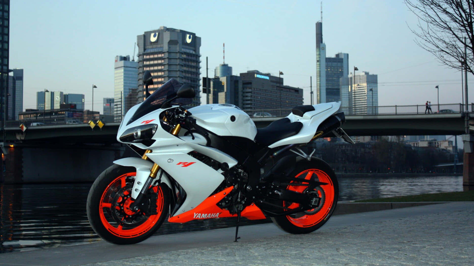  壁紙 — 一架白色和橙色的Yamaha運動電單車停泊在河邊小徑上，背景是黃昏時分的現代城市天際線。