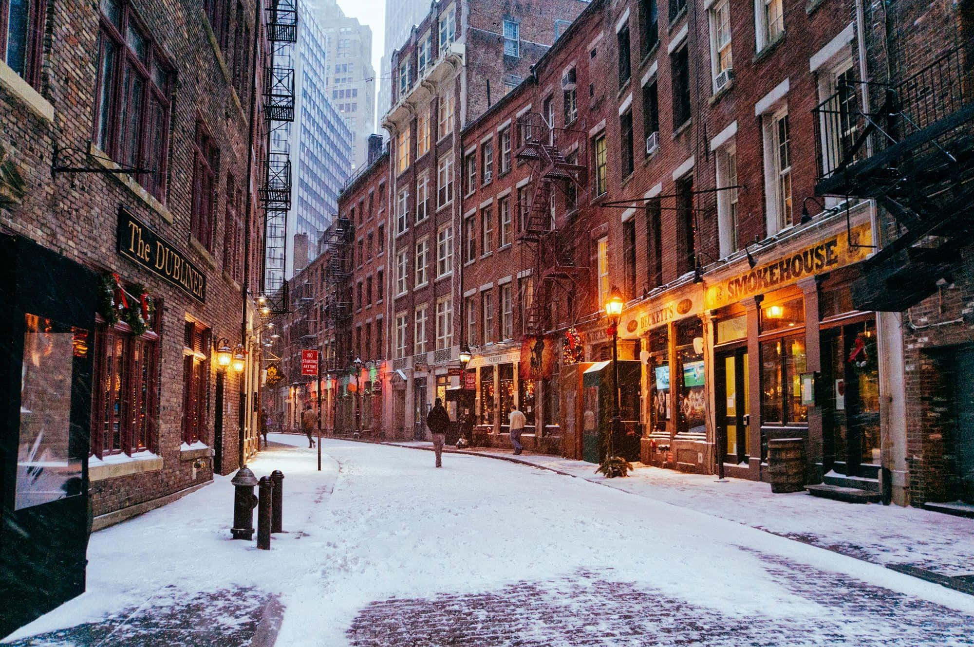 Winter Cityscape 桌布 壁紙 — 白雪覆蓋歷史建築之間狹窄的磚砌小巷，溫暖的燈光和節日裝飾閃爍。