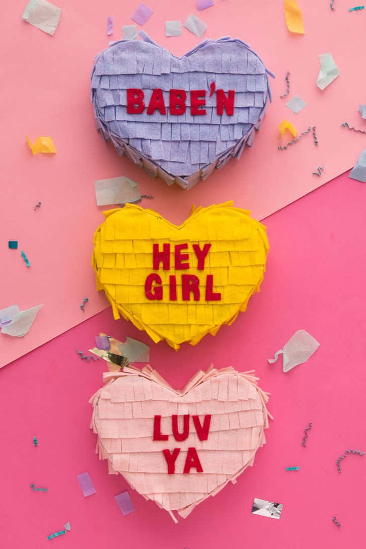 Babe'n Hey Girl Luv Ya情人節圖片 壁紙 — 三個色彩繽紛的心形皮納塔，帶有深情訊息，放在一個散落著五彩紙屑的充滿活力的粉紅色背景上。